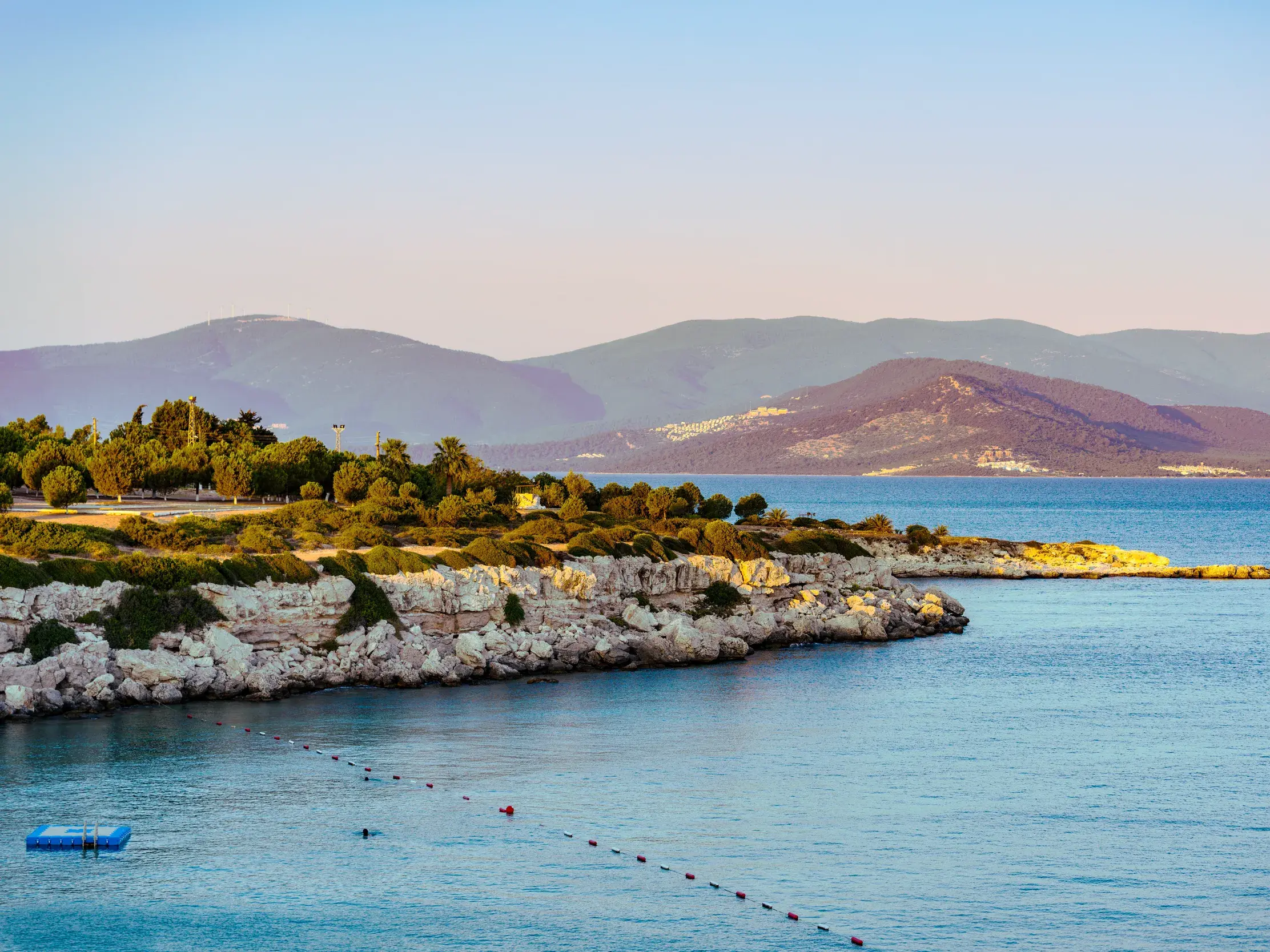 Idyllische Bucht an der Ägäis in der Türkei: Helle Felsklippen und ruhiges, blaues Meer vor einer Bergkulisse bei Kusadasi.
