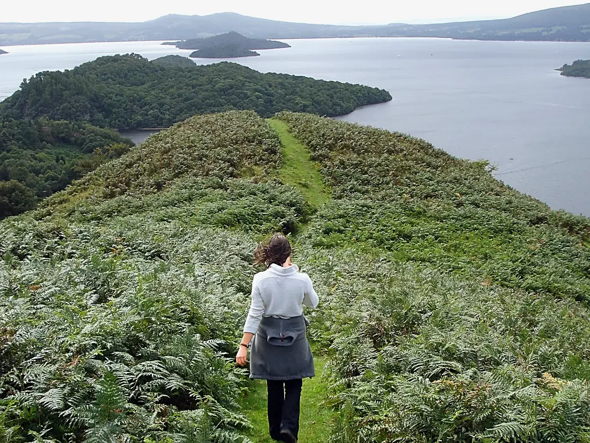Eine Frau wandert auf einem schmalen Pfad durch eine üppige Landschaft mit hohen Farnen in den schottischen Highlands.