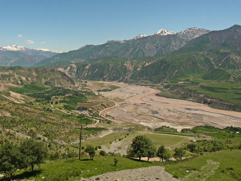 Weiter Blick über das grüne Rasht-Tal in Tadschikistan, durch das ein breiter Fluss fließt, hin zu den schneebedeckten Pamir-Gipfeln.
