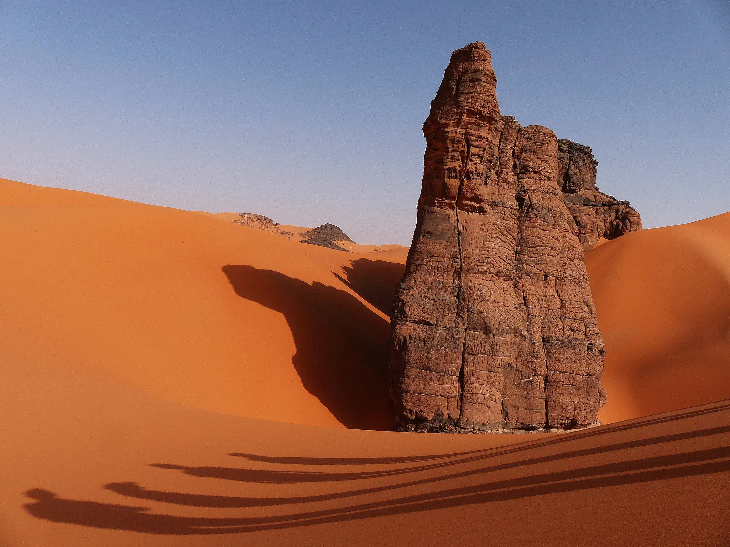 Ein markanter Felsturm ragt aus den leuchtend orangen Dünen im Tadrart Nationalpark in der Sahara Algeriens hervor.