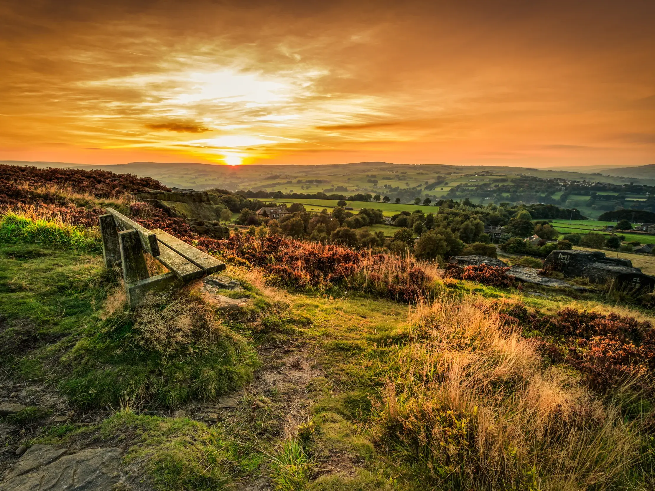 Eine Holzbank auf einer Anhöhe mit weitem Blick über Yorkshires hügelige Moorlandschaft bei Sonnenuntergang.