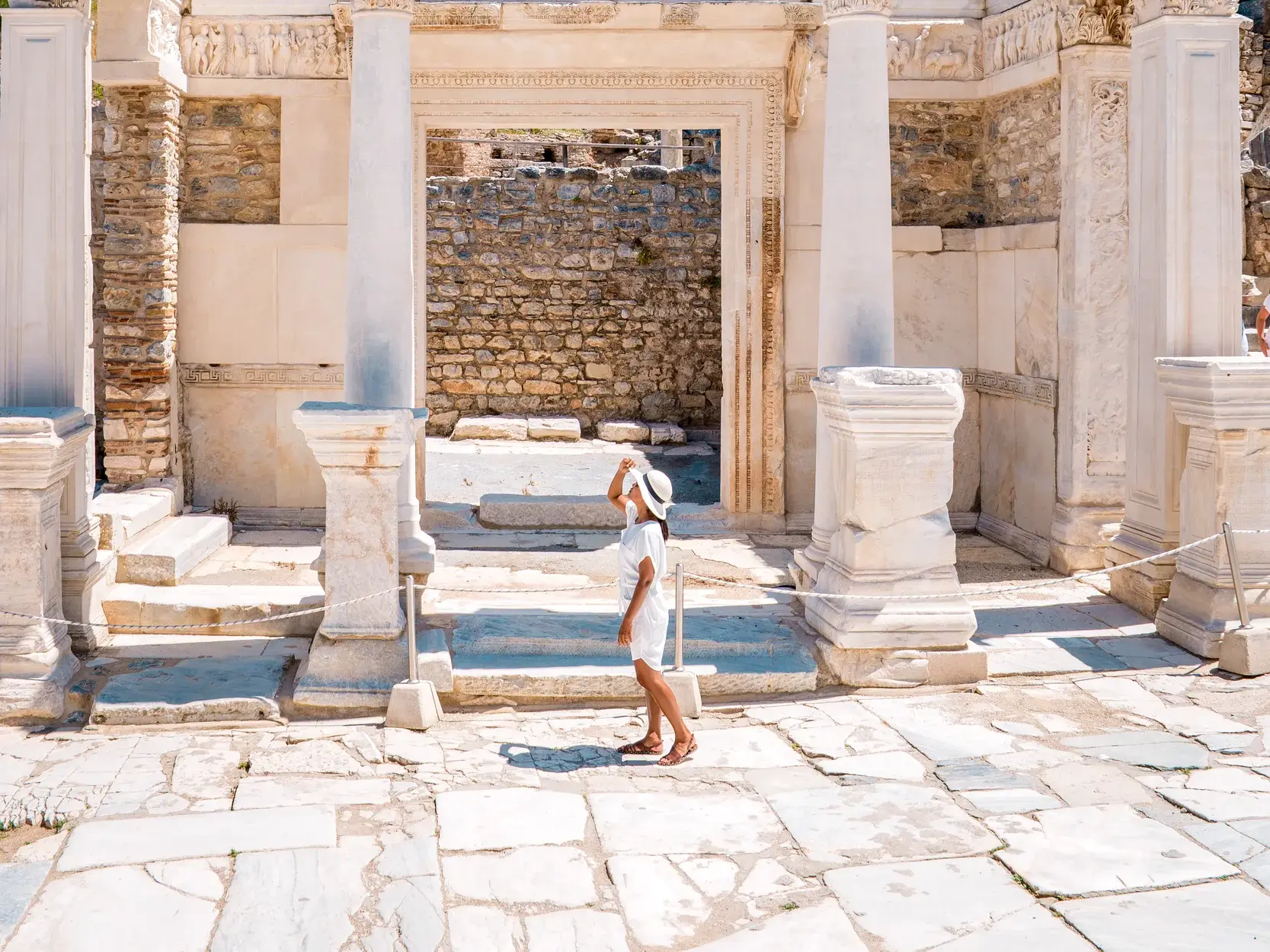 Eine Reisende mit weißem Hut blickt zur reich verzierten Fassade mit Säulen und Bögen in der antiken Stätte Ephesus, Türkei.