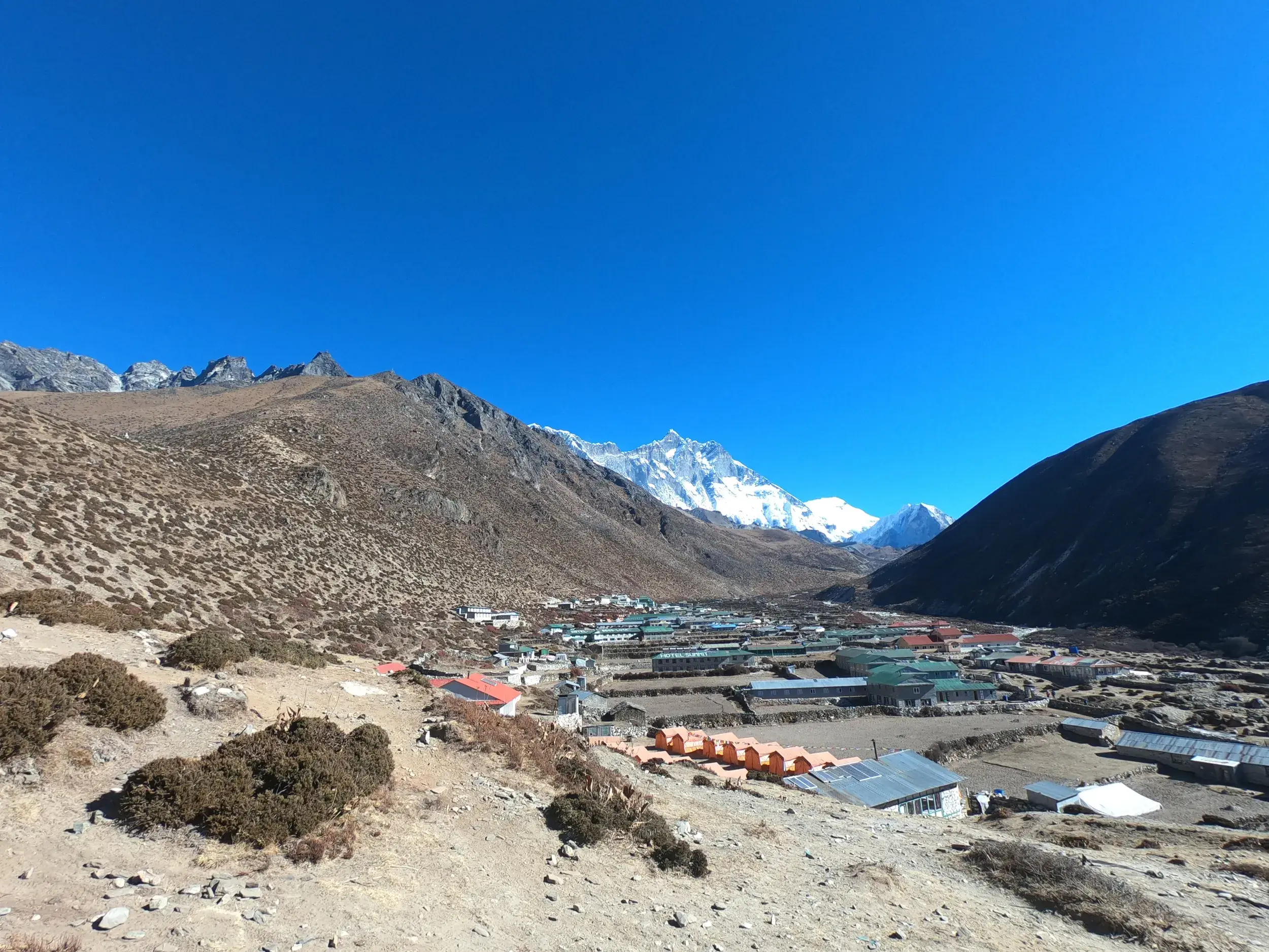 Blick von einem Höhenweg auf das Bergdorf Pheriche, eingebettet in ein Tal vor den schneebedeckten Eisriesen des Himalaya in Nepal.