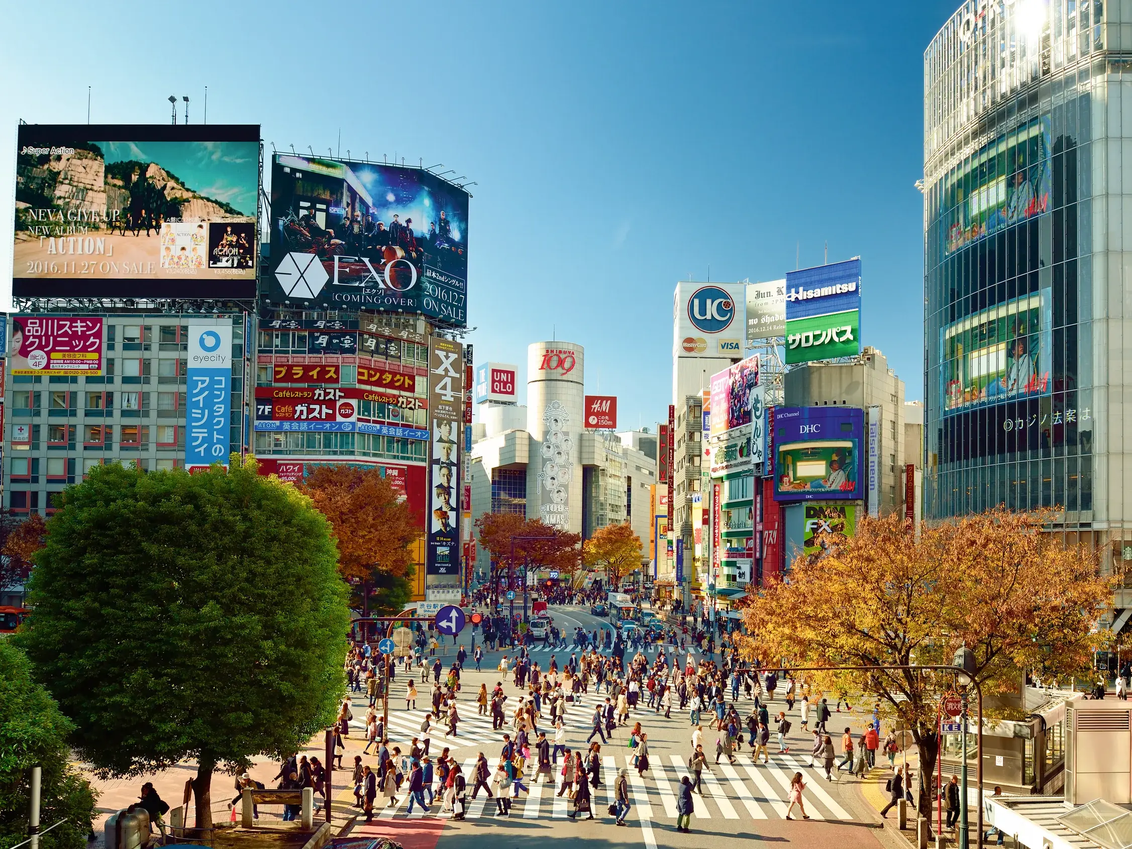 Hunderte Menschen überqueren die berühmte Shibuya Kreuzung in Tokio im Herbst, umgeben von Hochhäusern mit Neonreklamen.