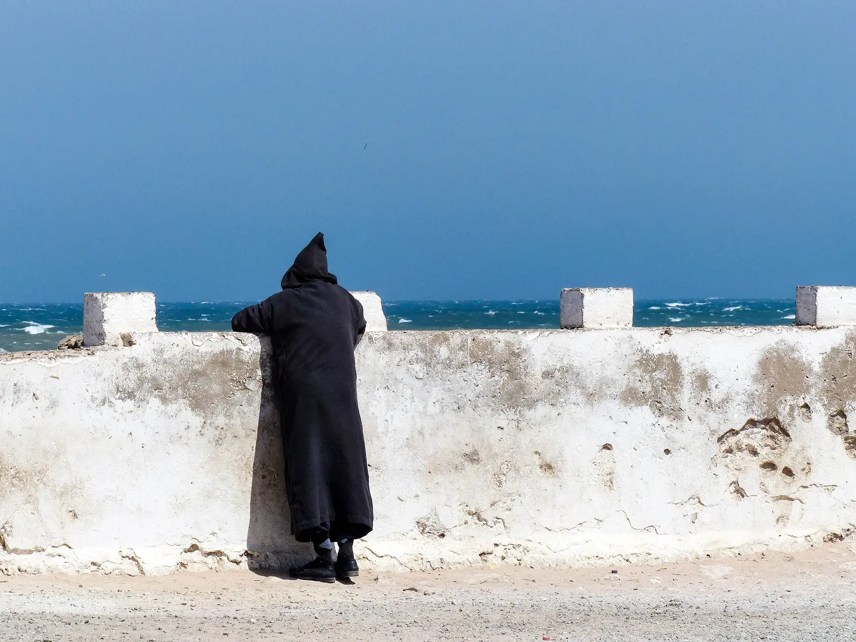 Einheimischer in schwarzer Djellaba blickt von der alten Stadtmauer in Essaouira auf den rauen, blauen Atlantik.