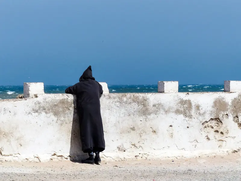 Einheimischer in schwarzer Djellaba blickt von der alten Stadtmauer in Essaouira auf den rauen, blauen Atlantik.