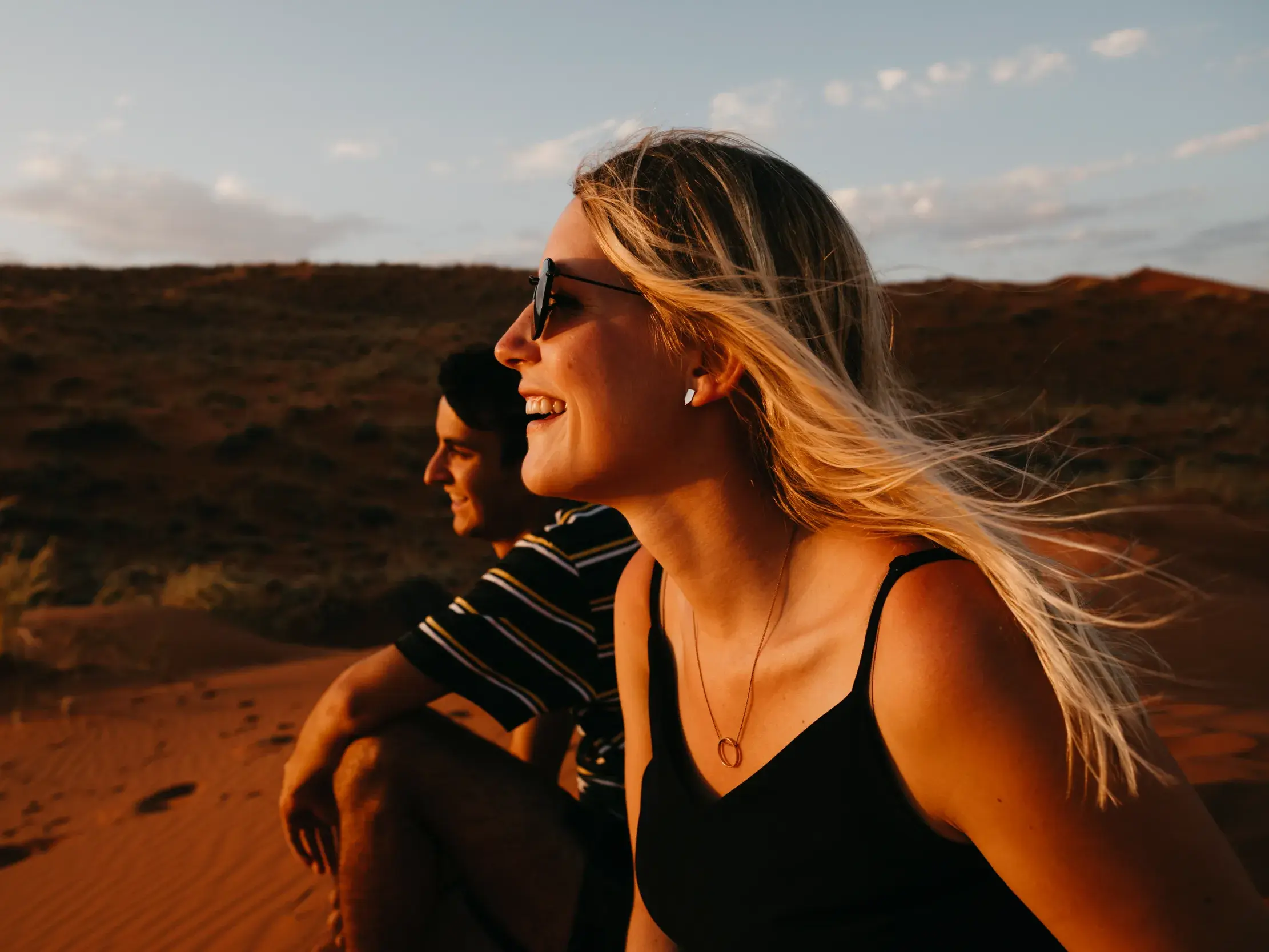 Eine junge Frau und ein Mann genießen den Sonnenuntergang auf einer roten Sanddüne in der Namib-Wüste während ihrer Gruppenreise.