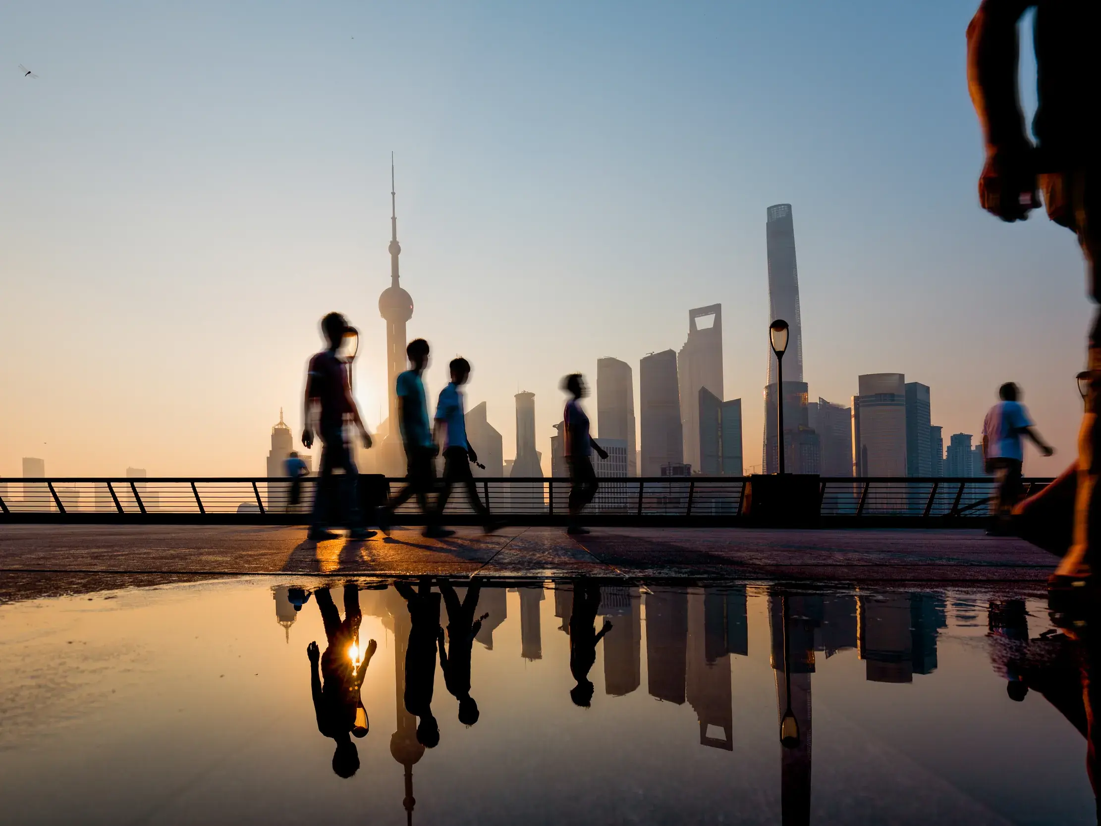Silhouetten von Passanten am Bund in Shanghai bei Sonnenaufgang, die Skyline von Pudong spiegelt sich in einer Wasserpfütze.