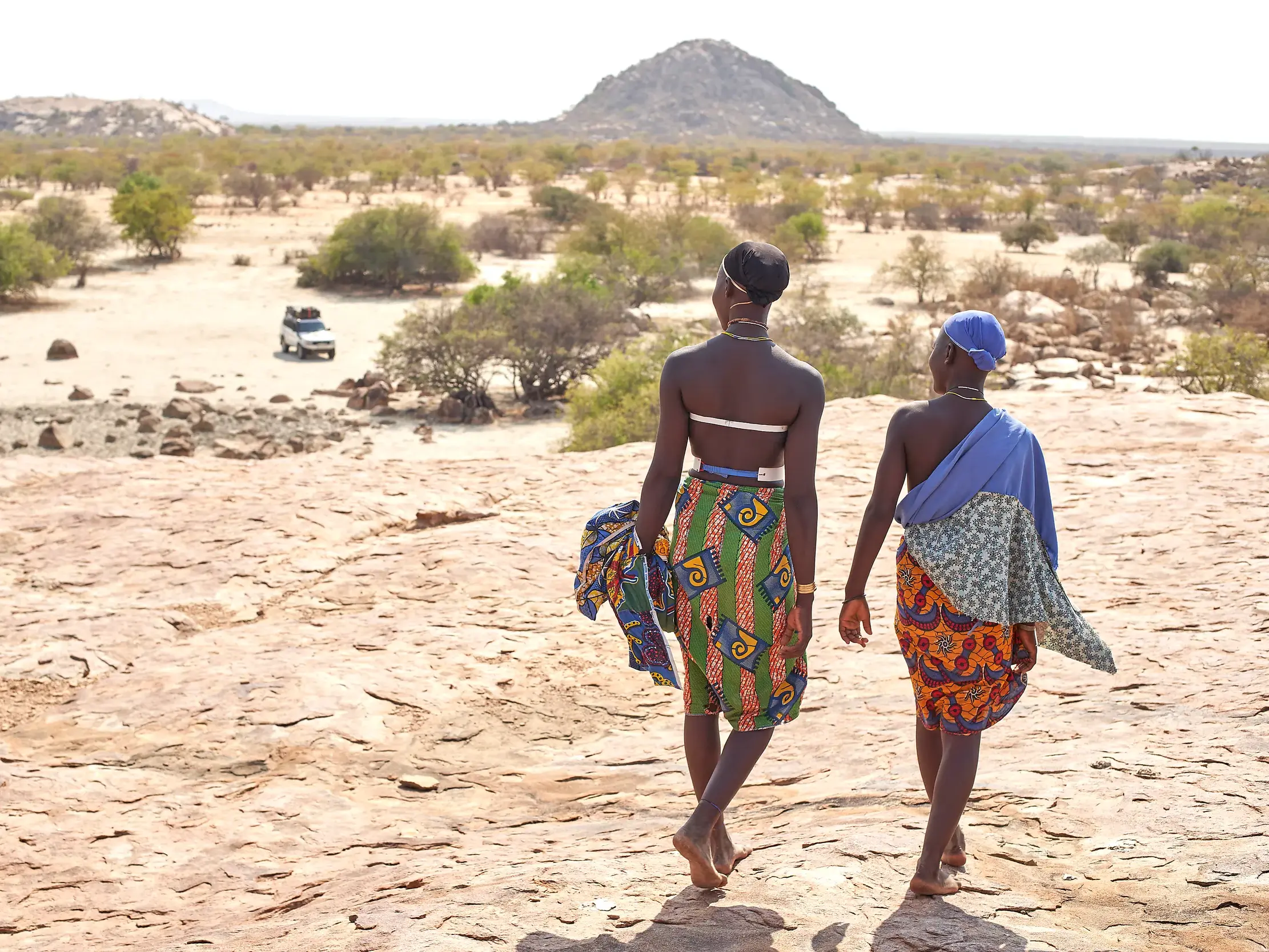 Zwei Frauen in bunten traditionellen Gewändern gehen barfuß durch die weite, felsige Landschaft im Süden von Angola.