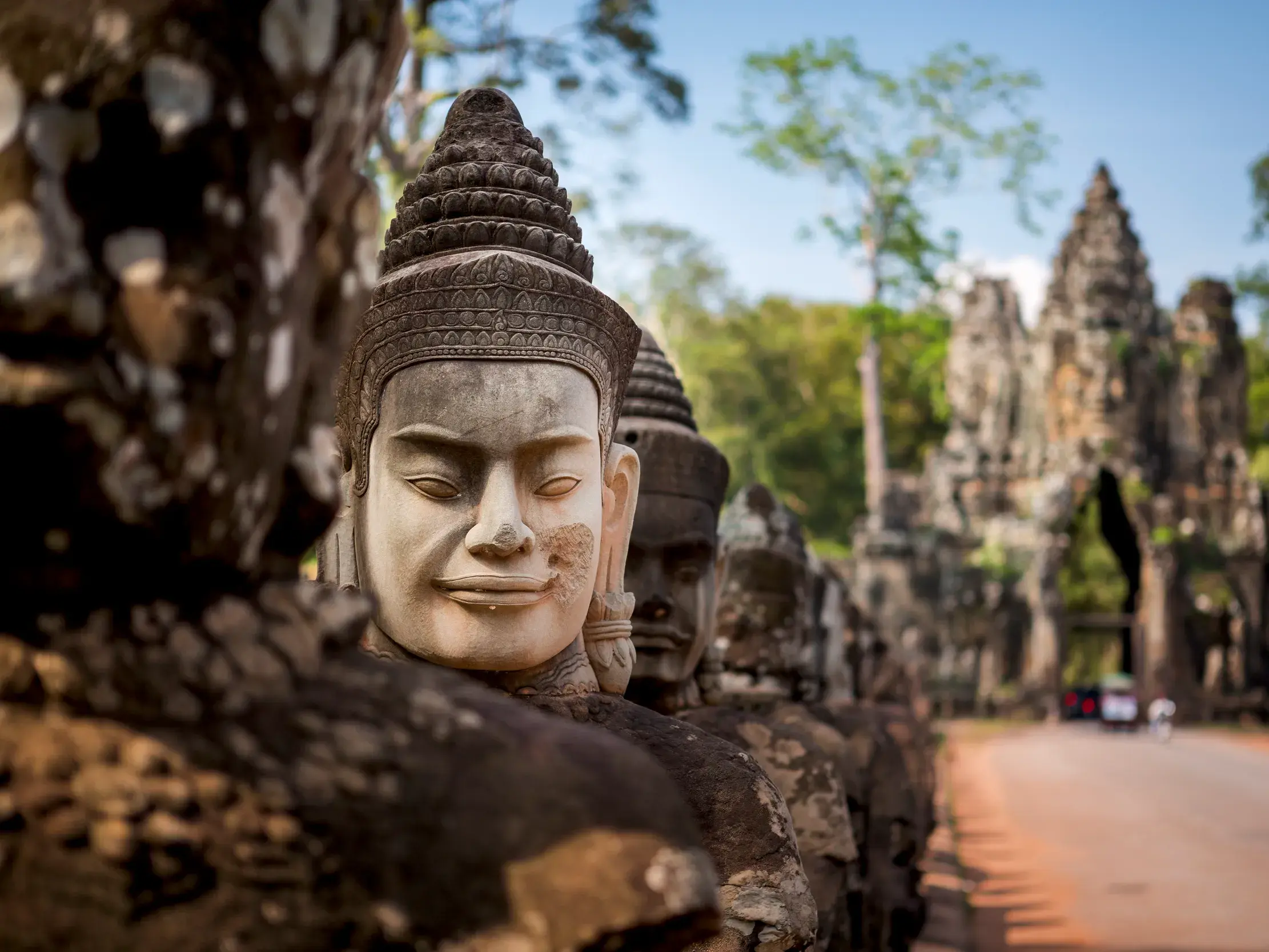Steinerne Götterfiguren säumen die Brücke zum Tor von Angkor Thom in Kambodscha, ein Tuk-Tuk nähert sich im Hintergrund.