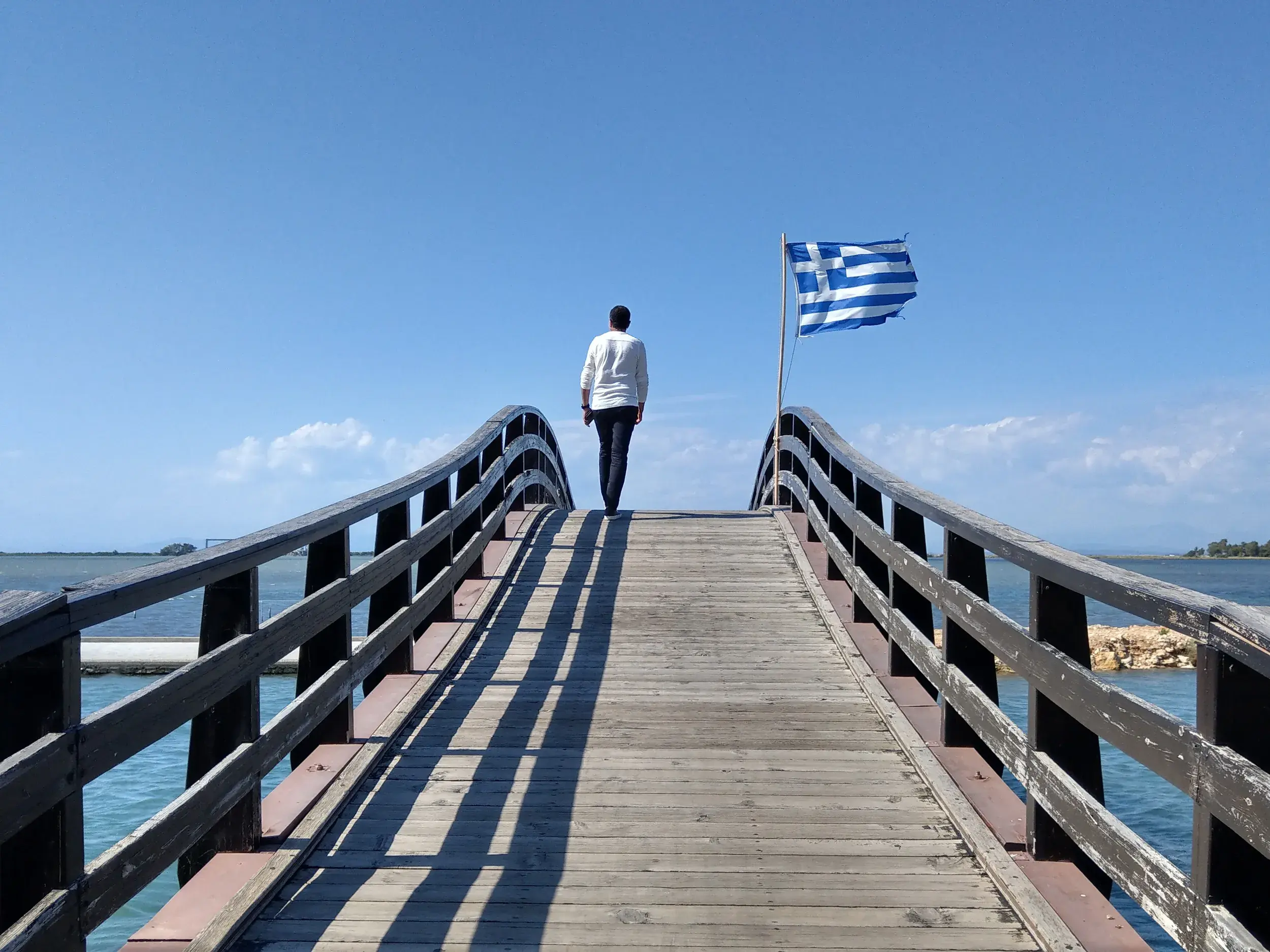 Ein Wanderer überquert eine hölzerne Bogenbrücke am Meer in Griechenland, während neben ihm eine griechische Flagge weht.
