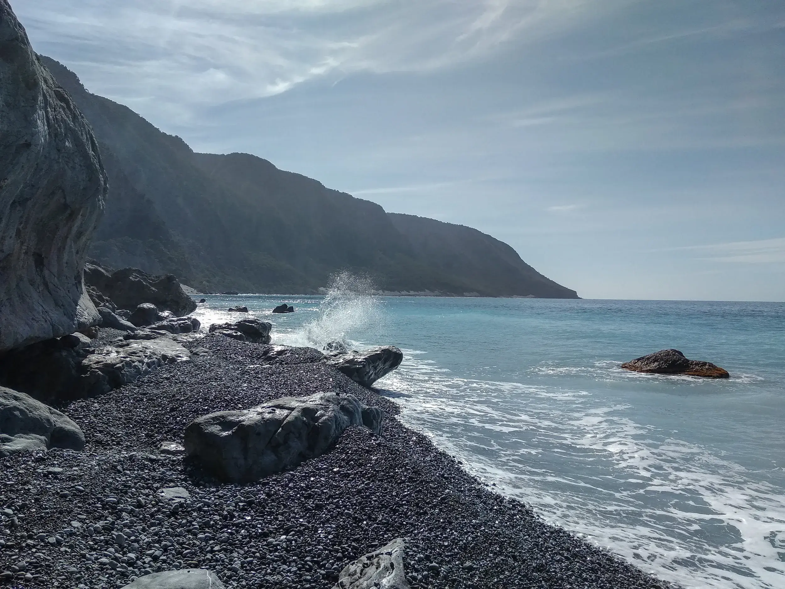 Eine Welle bricht an einem einsamen Kieselstrand an Kretas wilder Südküste, im Hintergrund die Ausläufer der Lefka Ori Berge.