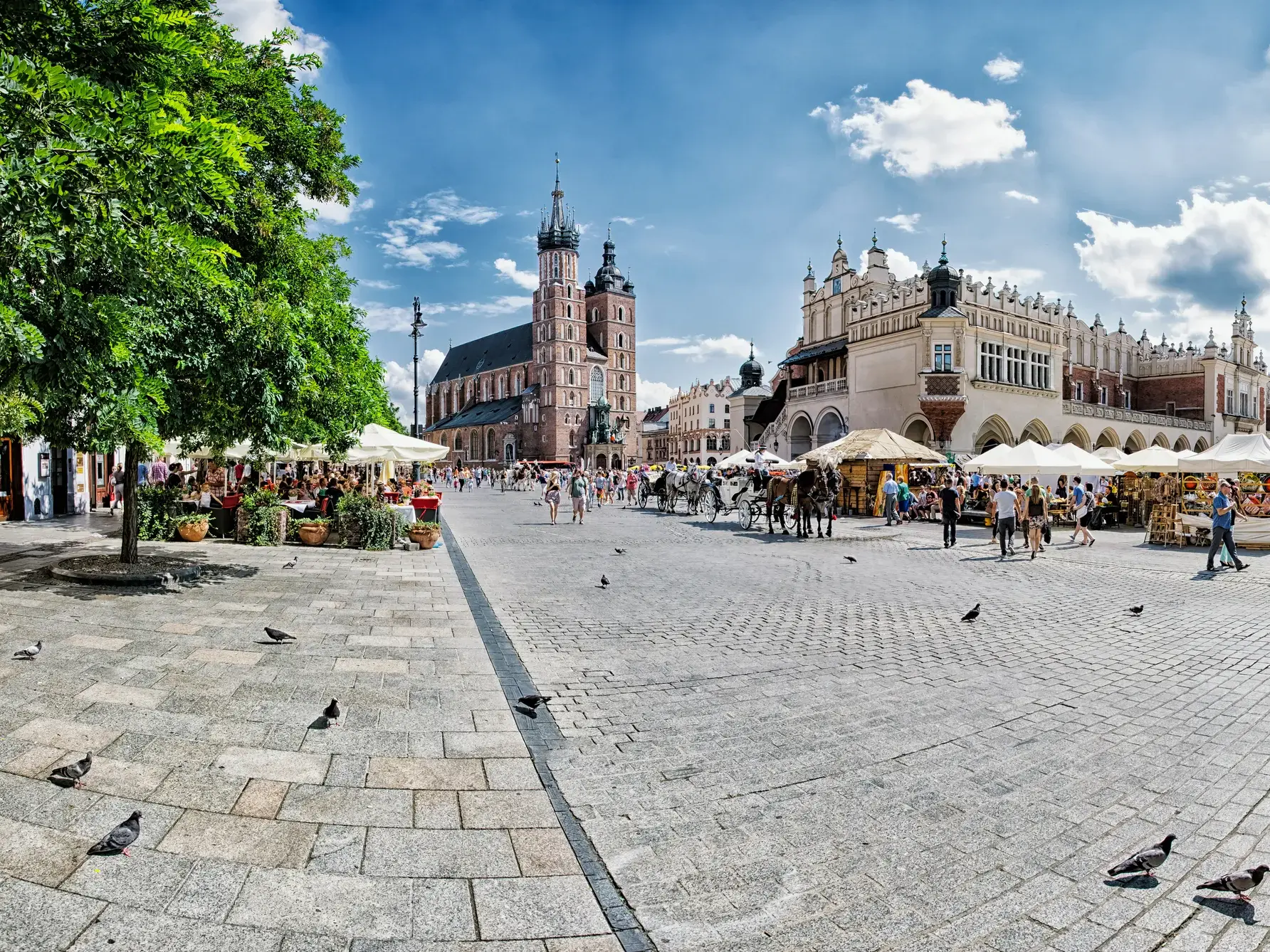 Touristen flanieren bei Sonnenschein über den Marktplatz der Altstadt von Krakau, vorbei an den Tuchhallen und der Marienkirche.