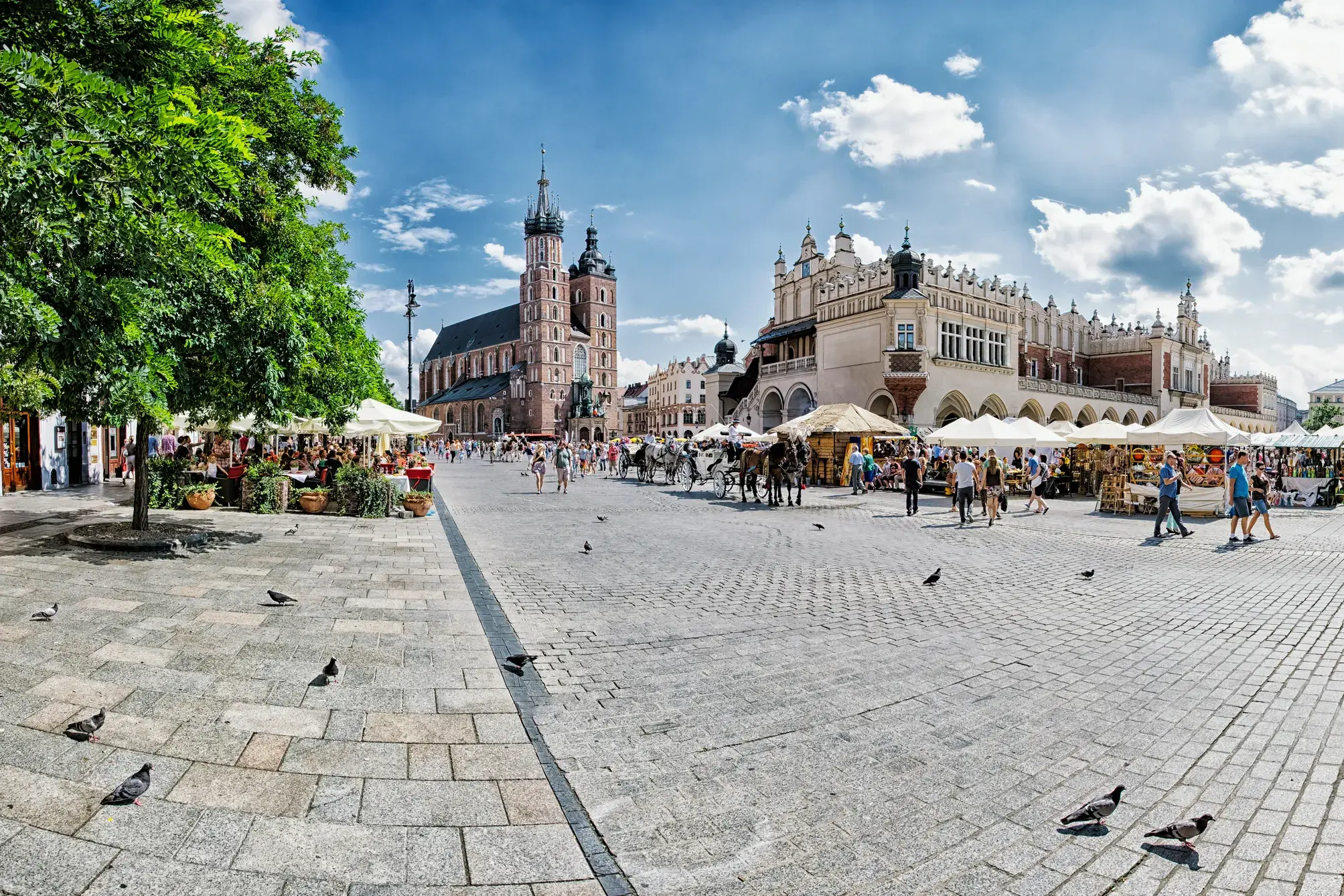 Touristen flanieren bei Sonnenschein über den Marktplatz der Altstadt von Krakau, vorbei an den Tuchhallen und der Marienkirche.