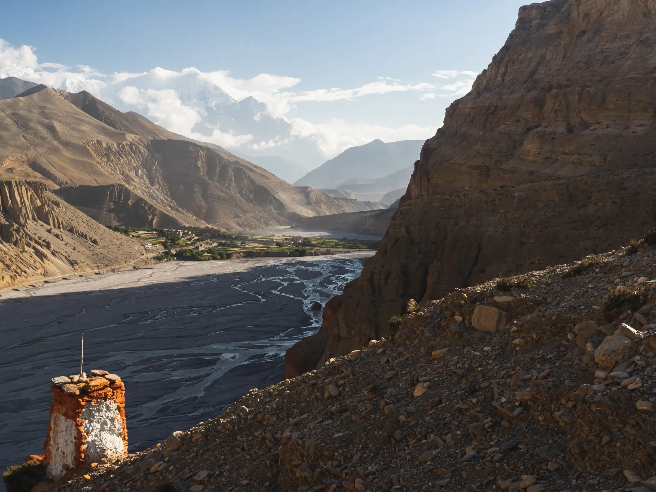 Weite Aussicht beim Trekking in Mustang über das Kali Gandaki-Tal mit einem Dorf und der schneebedeckten Annapurna-Kette am Horizont.