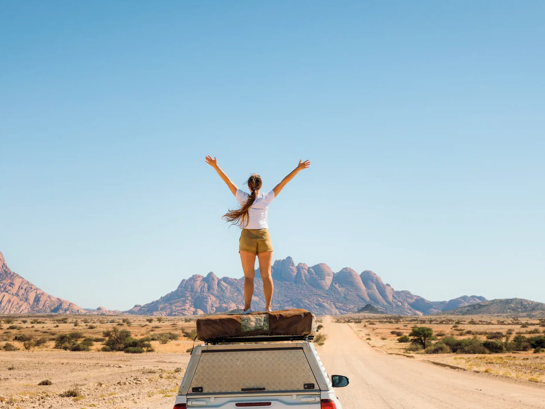 Junge Frau auf einer Rundreise durch Namibia, die jubelnd auf dem Dach eines Safari-Lkw vor der Spitzkoppe steht.