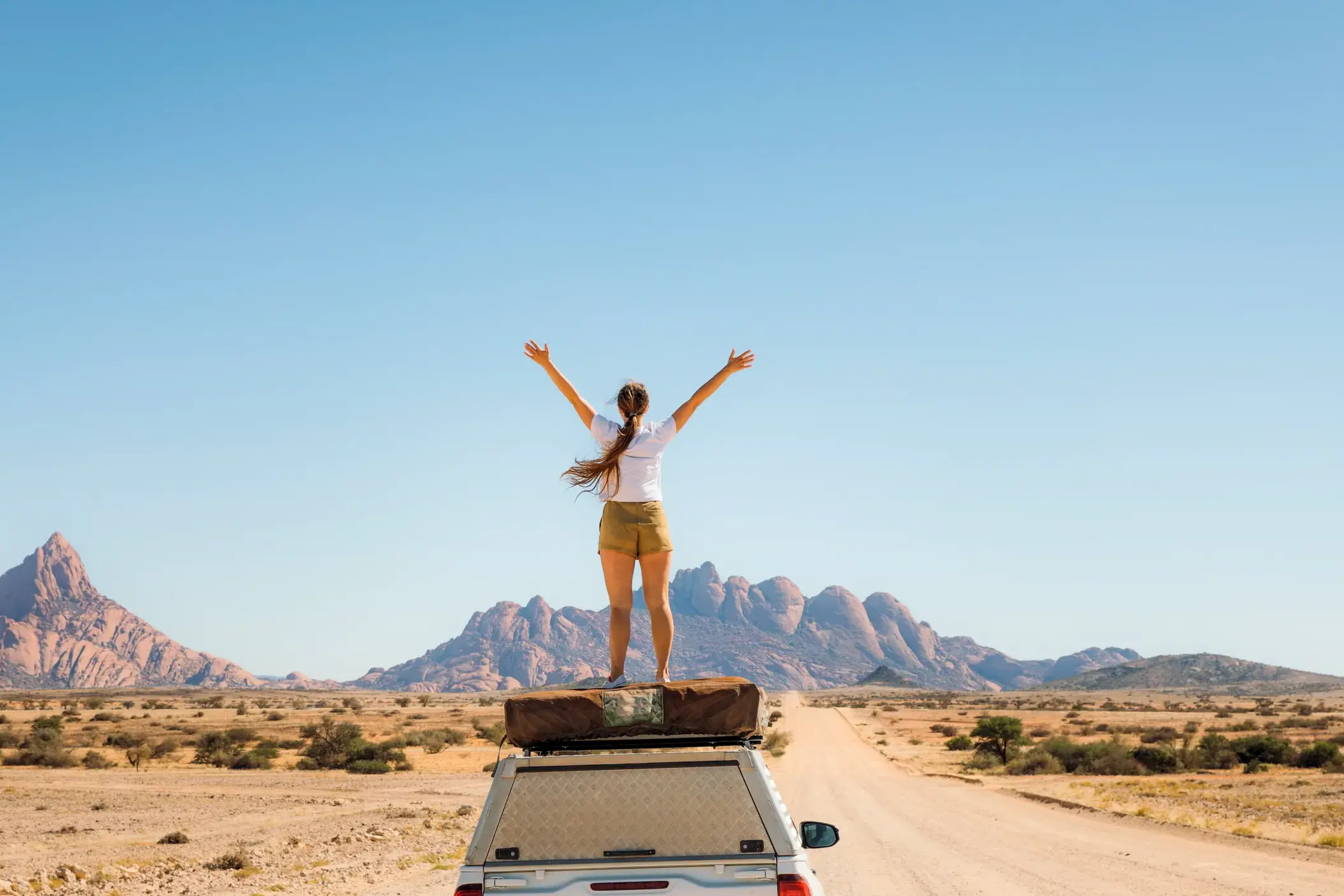 Junge Frau auf einer Rundreise durch Namibia, die jubelnd auf dem Dach eines Safari-Lkw vor der Spitzkoppe steht.
