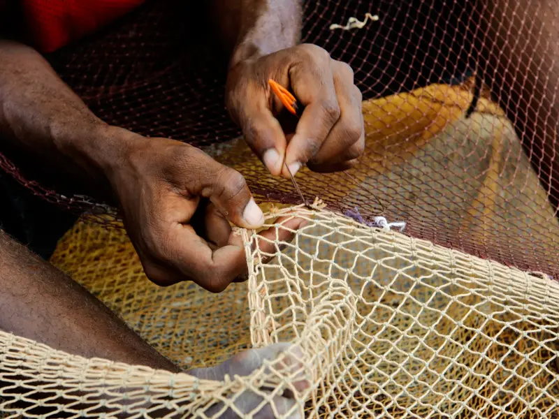 Nahaufnahme der Hände eines Fischers auf den Kapverden, der mit einem Netzknüpfer geschickt ein beigefarbenes Fischernetz repariert.