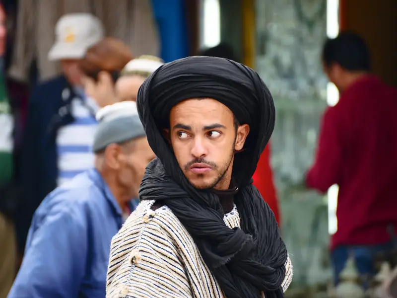 Ein junger Beduine als Reisebegleiter mit schwarzem Turban beobachtet das Geschehen auf einem belebten Markt in Marokko.
