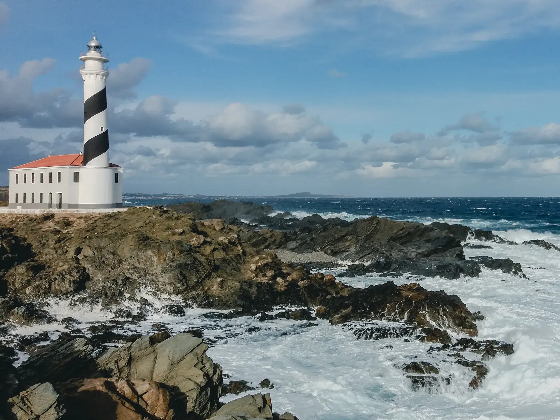 Wildes Mittelmeer brandet an die Felsküste am Cap de Favàritx mit seinem schwarz-weiß gestreiften Leuchtturm auf Menorca.