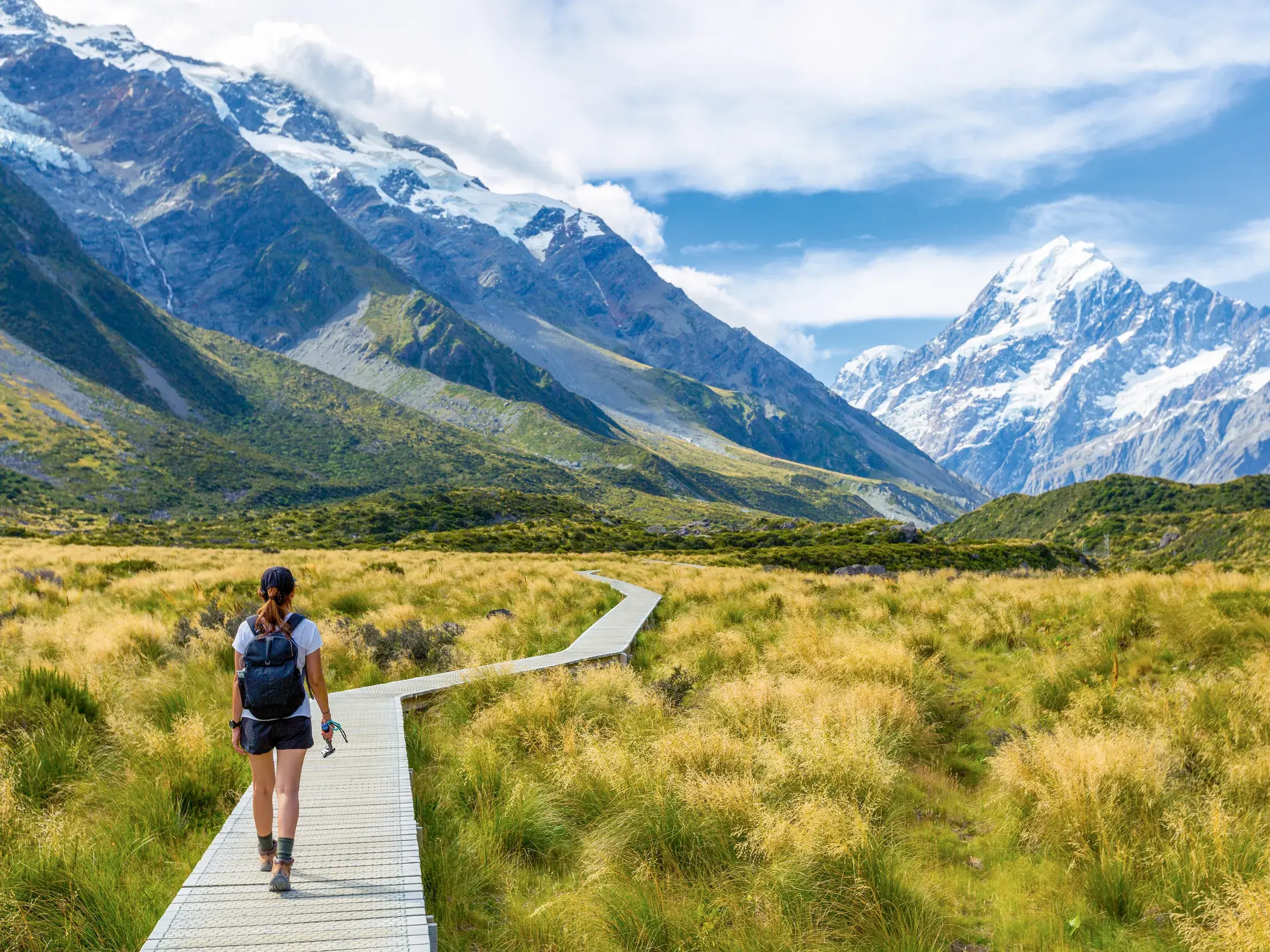 Eine Wanderin auf dem Holzsteg des Hooker Valley Tracks in Neuseeland mit Blick auf das schneebedeckte Mount Cook Massiv.