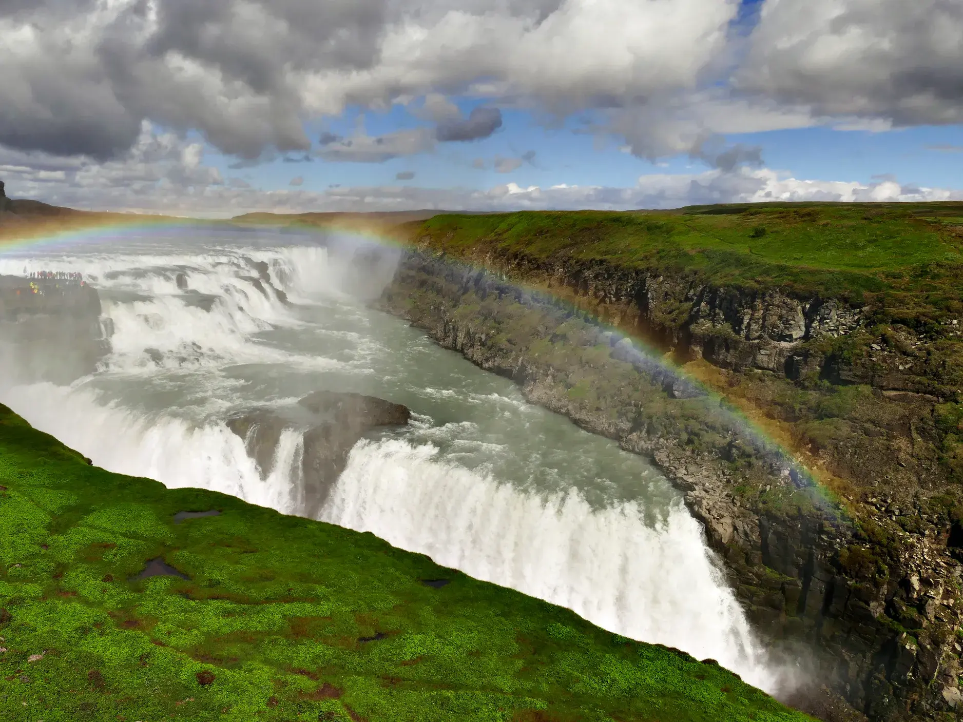 Ein leuchtender Regenbogen wölbt sich über den tosenden Gullfoss Wasserfall, der in eine moosgrüne Schlucht in Island stürzt.