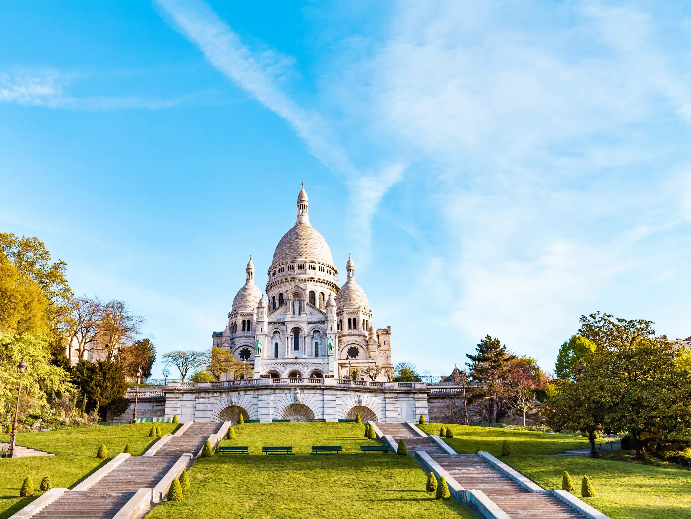 Die Basilika Sacré-Coeur auf dem Montmartre in Paris mit der grünen Parkanlage und den Treppen im Vordergrund bei blauem Himmel.
