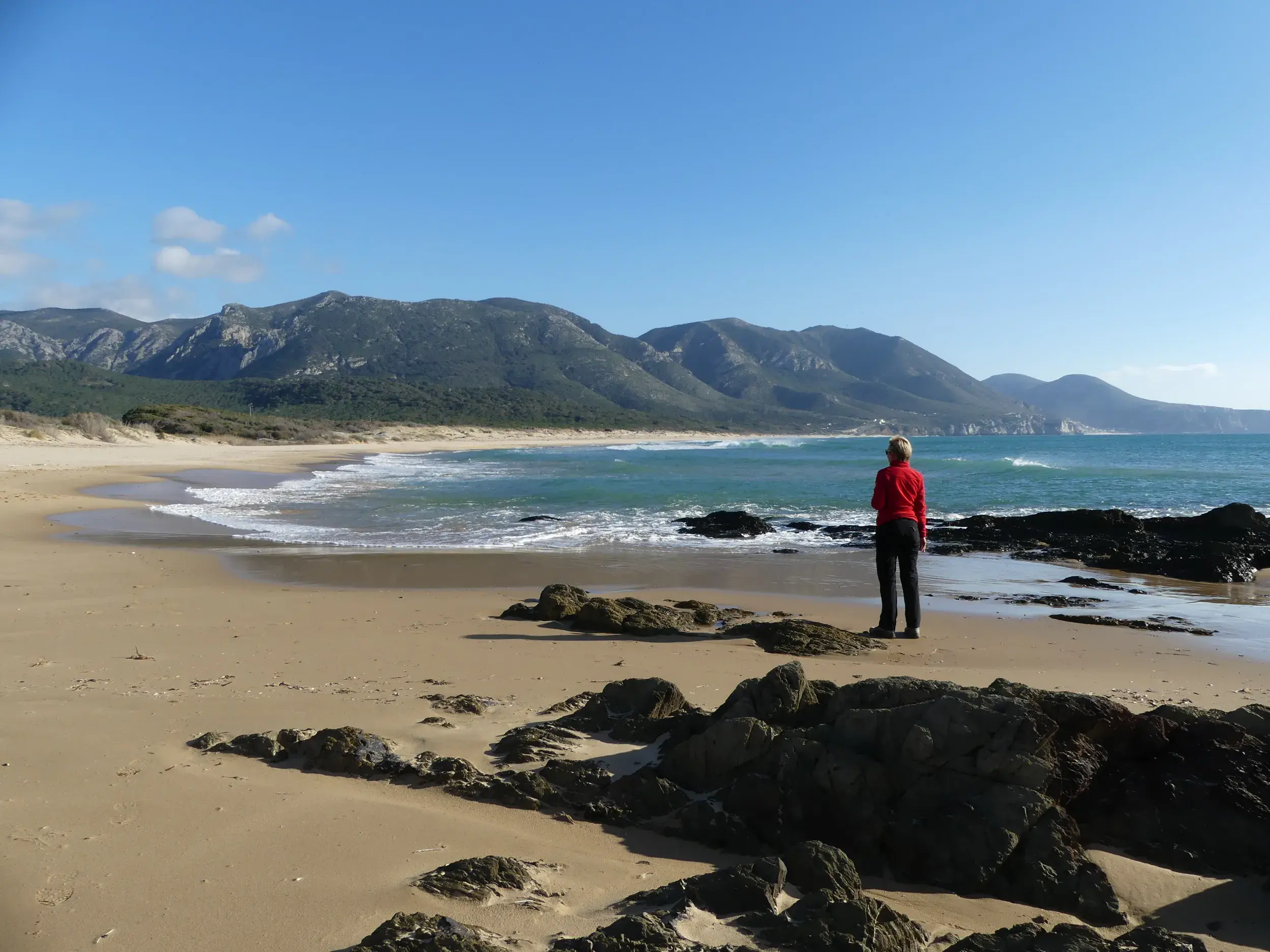 Wanderin am Sandstrand von Scivu an der Costa Verde auf Sardinien, blickt auf das Meer und die grüne Berglandschaft.