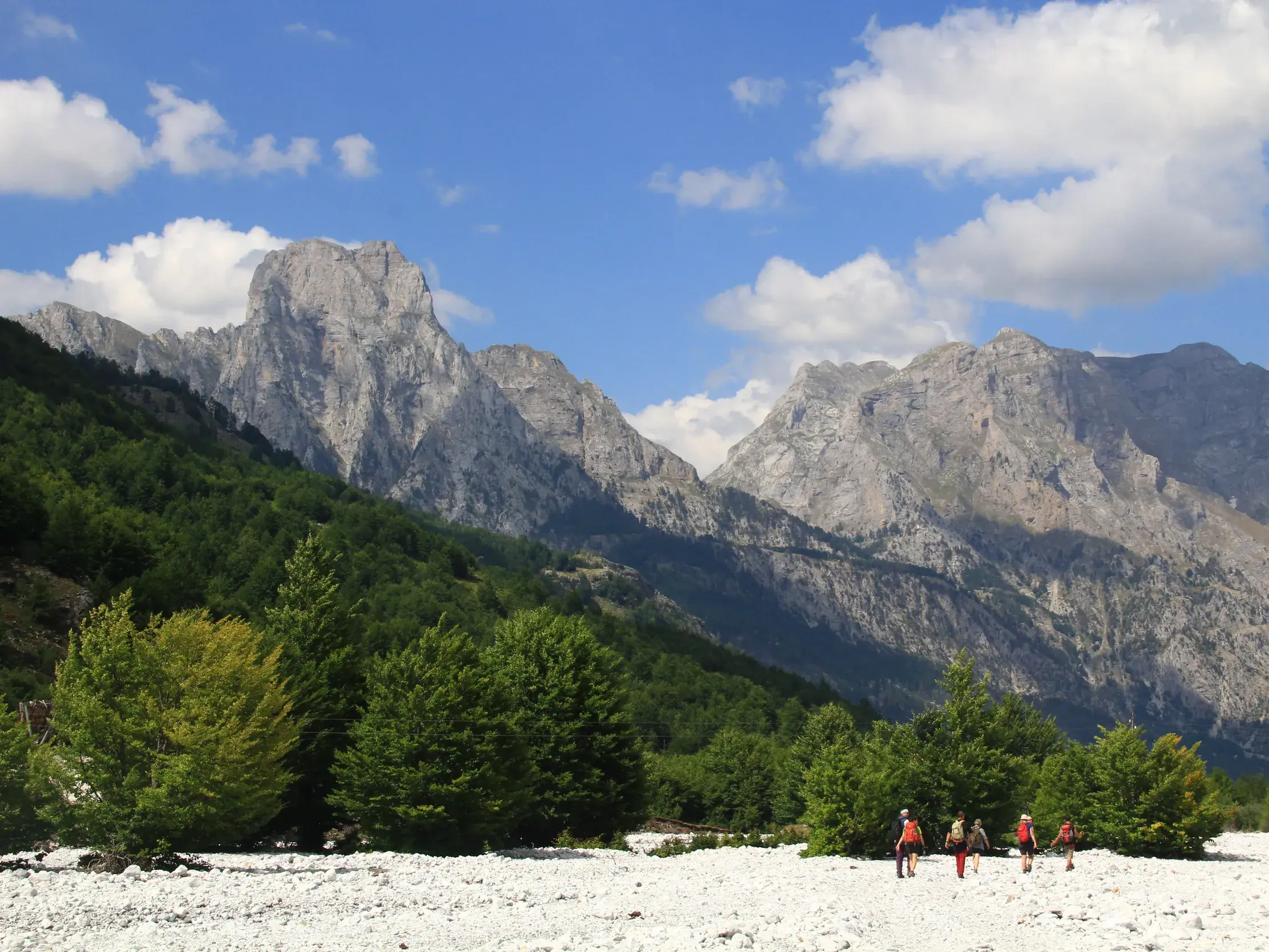 Eine kleine Wandergruppe durchquert ein weites, steiniges Flussbett im Valbona-Nationalpark vor beeindruckenden Berggipfeln.