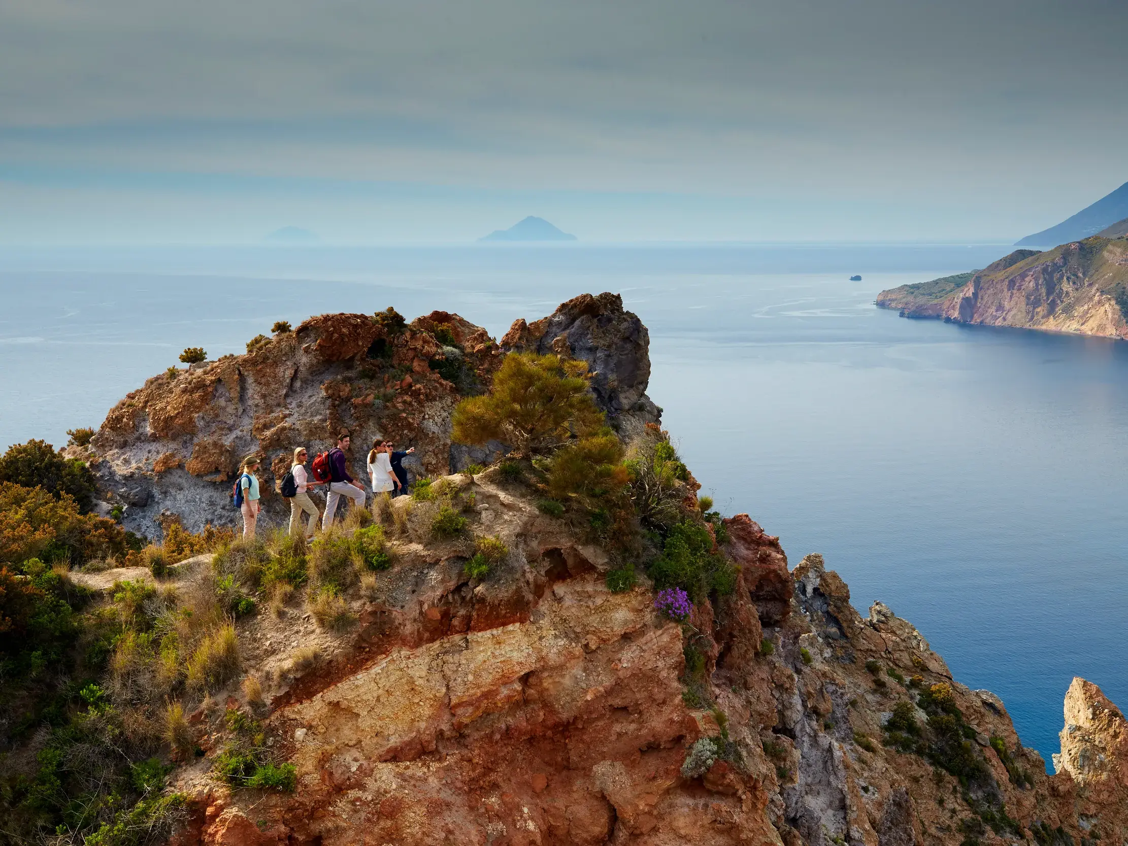 Eine kleine Wandergruppe auf einem Felsvorsprung genießt den weiten Ausblick auf die Küste der Äolischen Inseln Siziliens.