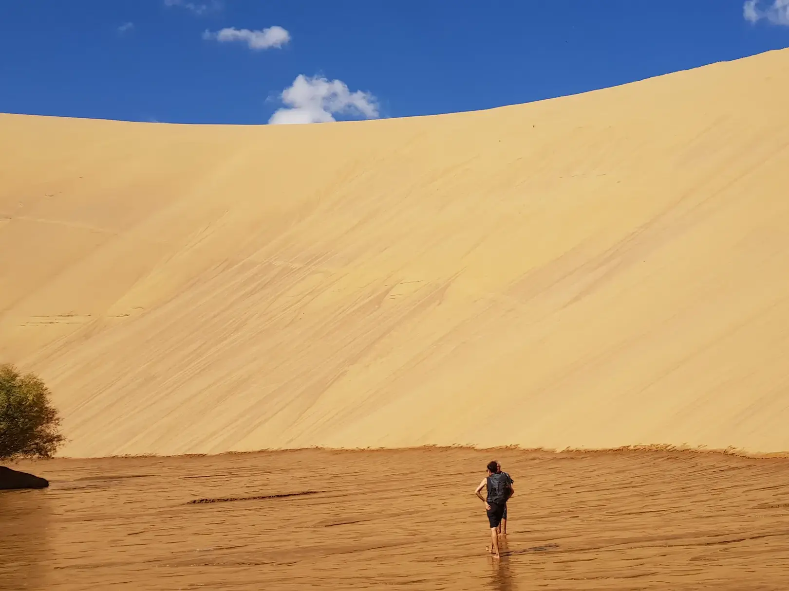 Ein Wanderer durchquert barfuß den Zavkhan Fluss vor der beeindruckenden Sanddüne von Mongol Els in der Mongolei.