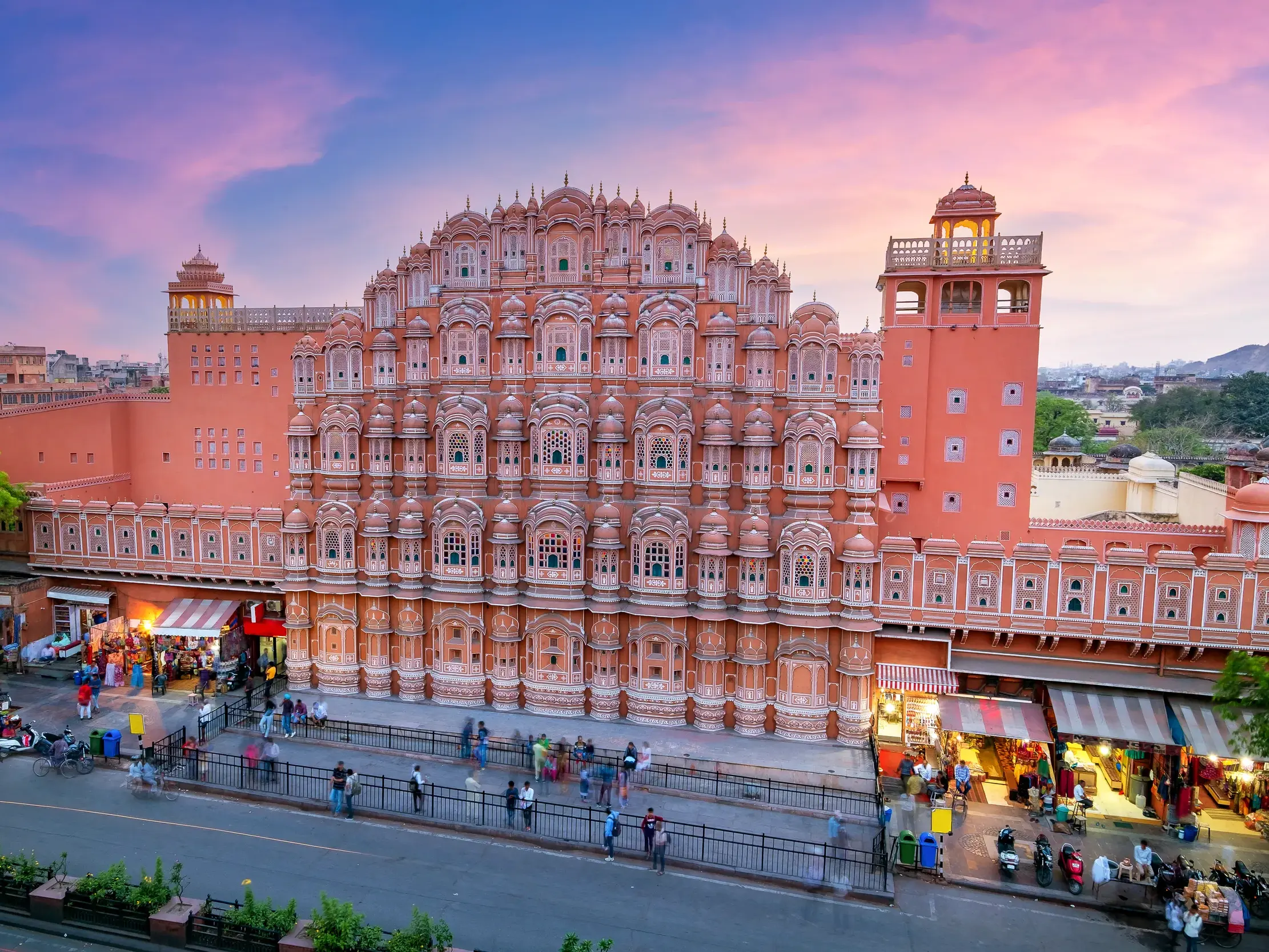 Die prachtvolle, rosarote Fassade des Hawa Mahal in Jaipur im Licht der Abenddämmerung über einer belebten Straße.