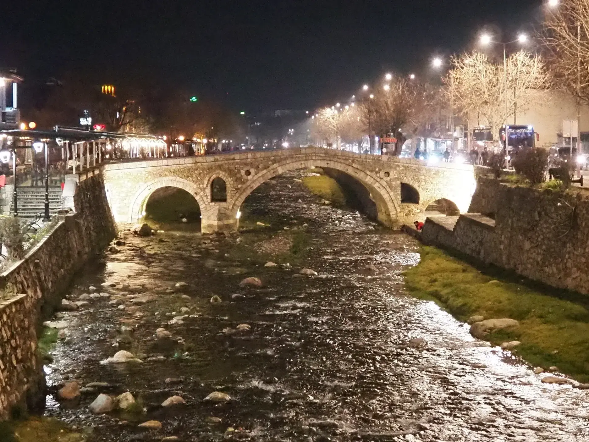Die historische Steinbrücke von Prizren bei Nacht, beleuchtet über den Fluss Lumbardhi, mit belebter Uferpromenade.
