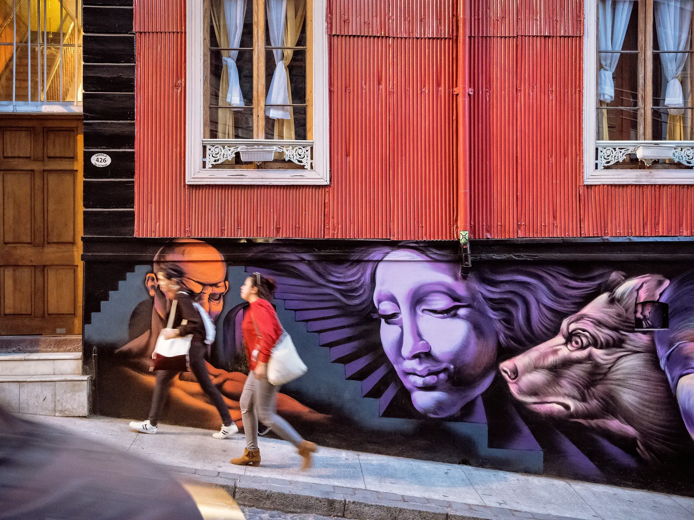 Passantinnen auf einem Gehweg in Valparaíso vor einer roten Wellblechfassade mit einem großen, surrealistischen Wandgemälde.