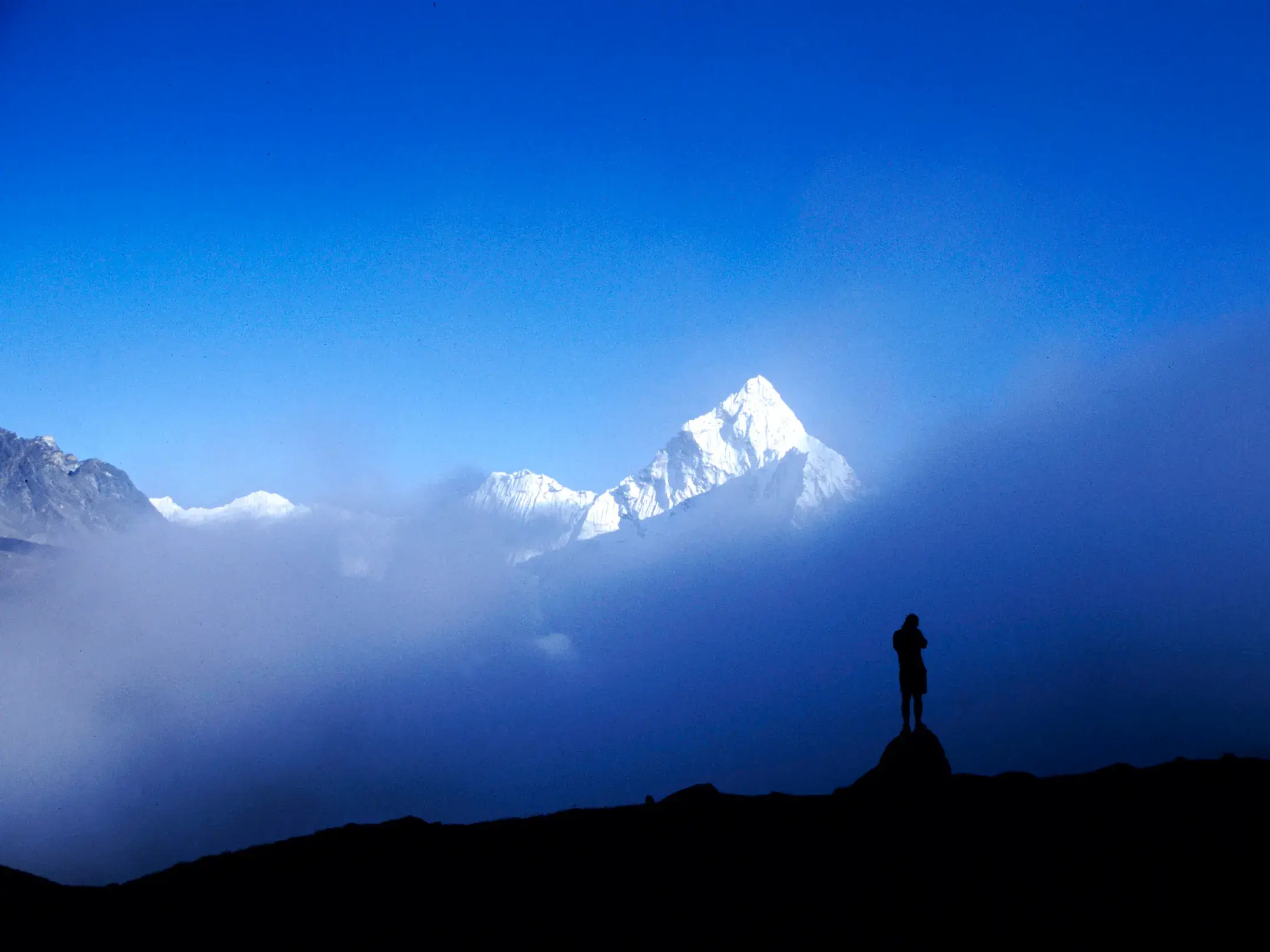 Ein Trekker auf einem Aussichtspunkt im Himalaya blickt auf einen Bergriesen, der aus einem Meer aus Wolken ragt.