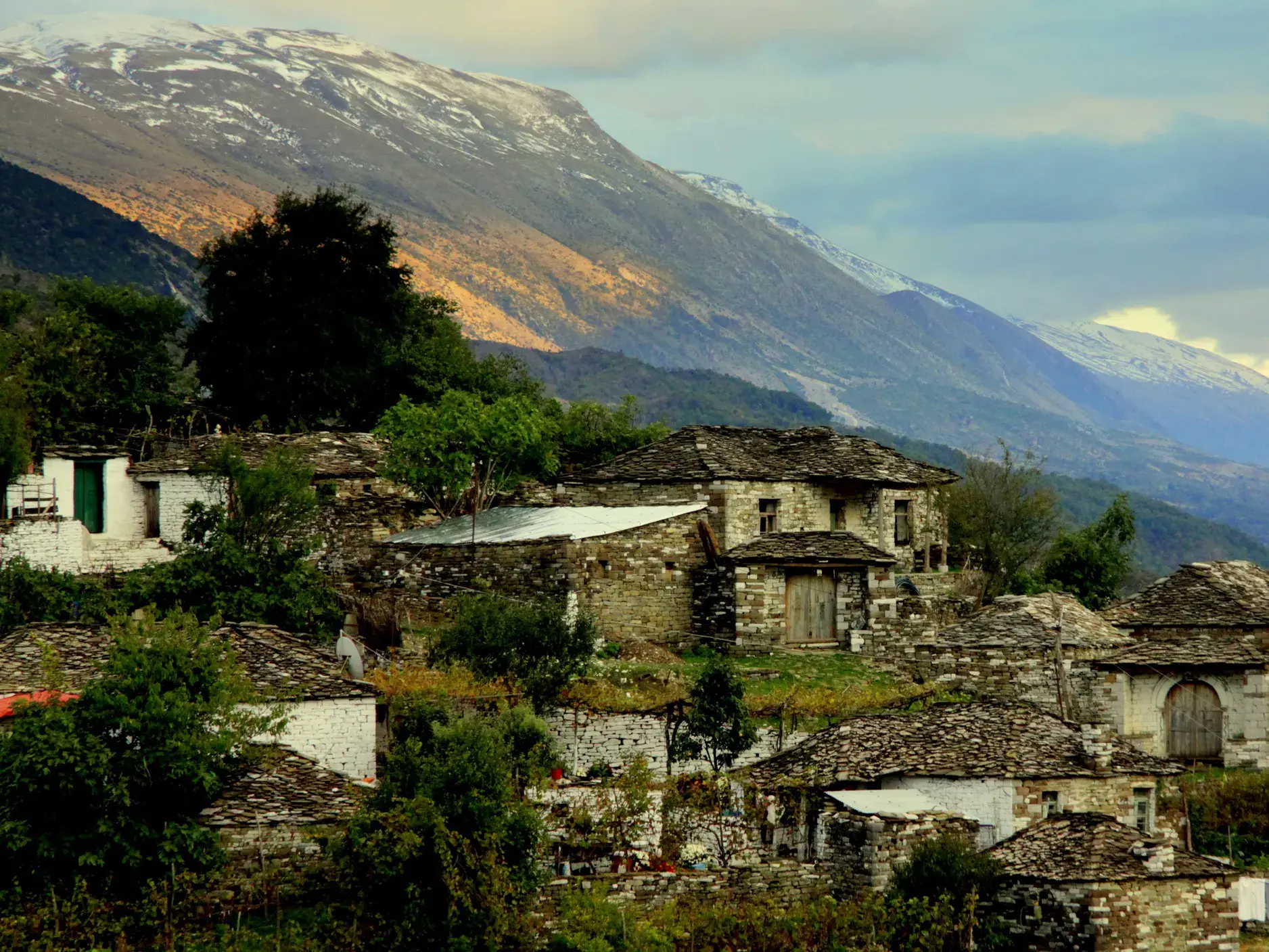 Traditionelle Steinhäuser eines Bergdorfs im Zagoria-Tal in Albanien, im Hintergrund ein schneebedeckter Berggipfel.
