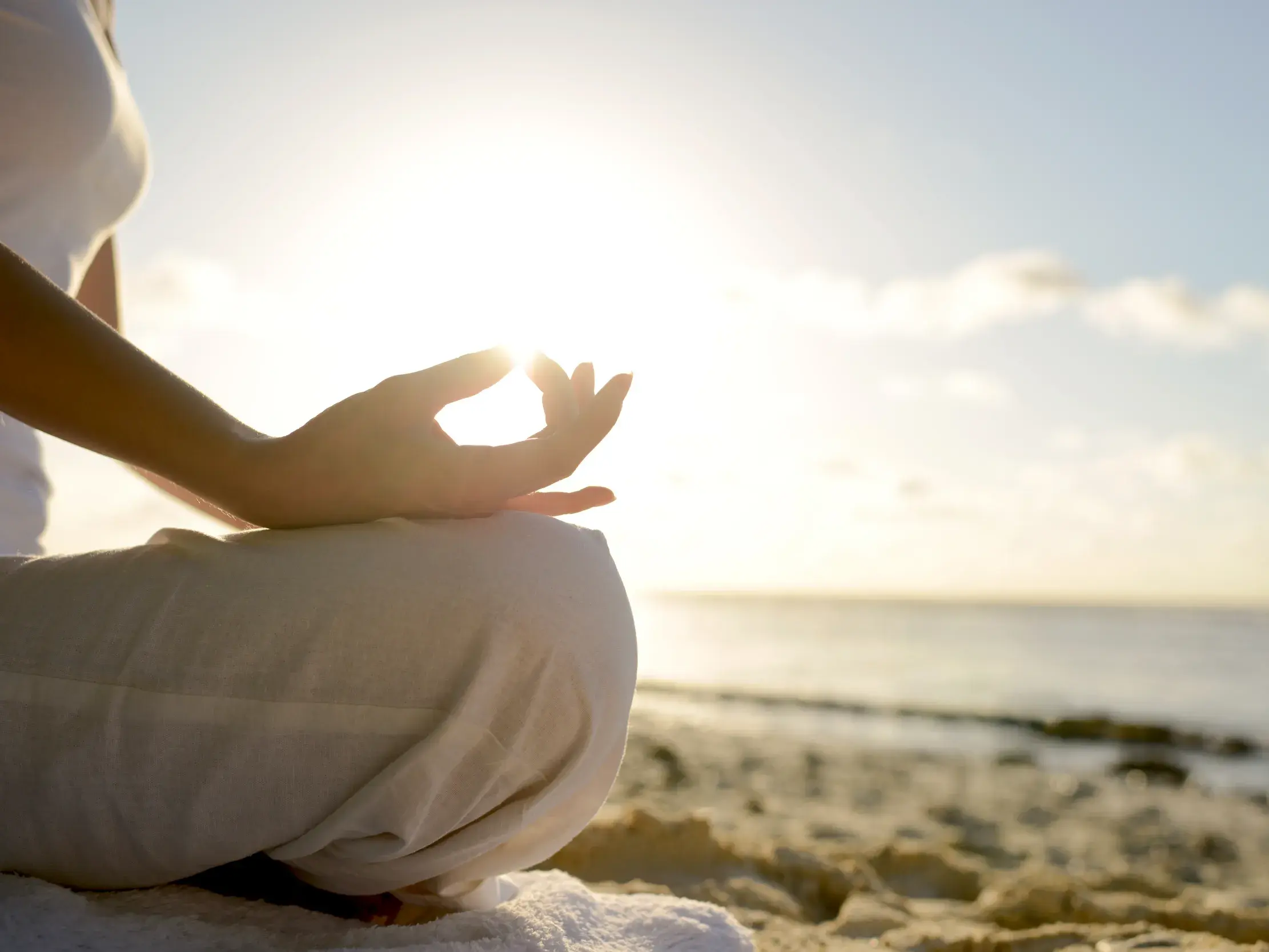 Eine Person praktiziert Yoga im Lotussitz am Strand in der Karibik, während die Sonne durch die Finger einer Hand scheint.