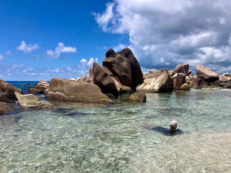 Ein Reiseteilnehmer badet im kristallklaren Wasser vor der Kulisse der einzigartigen Granitfelsen an einem Strand der Seychellen.