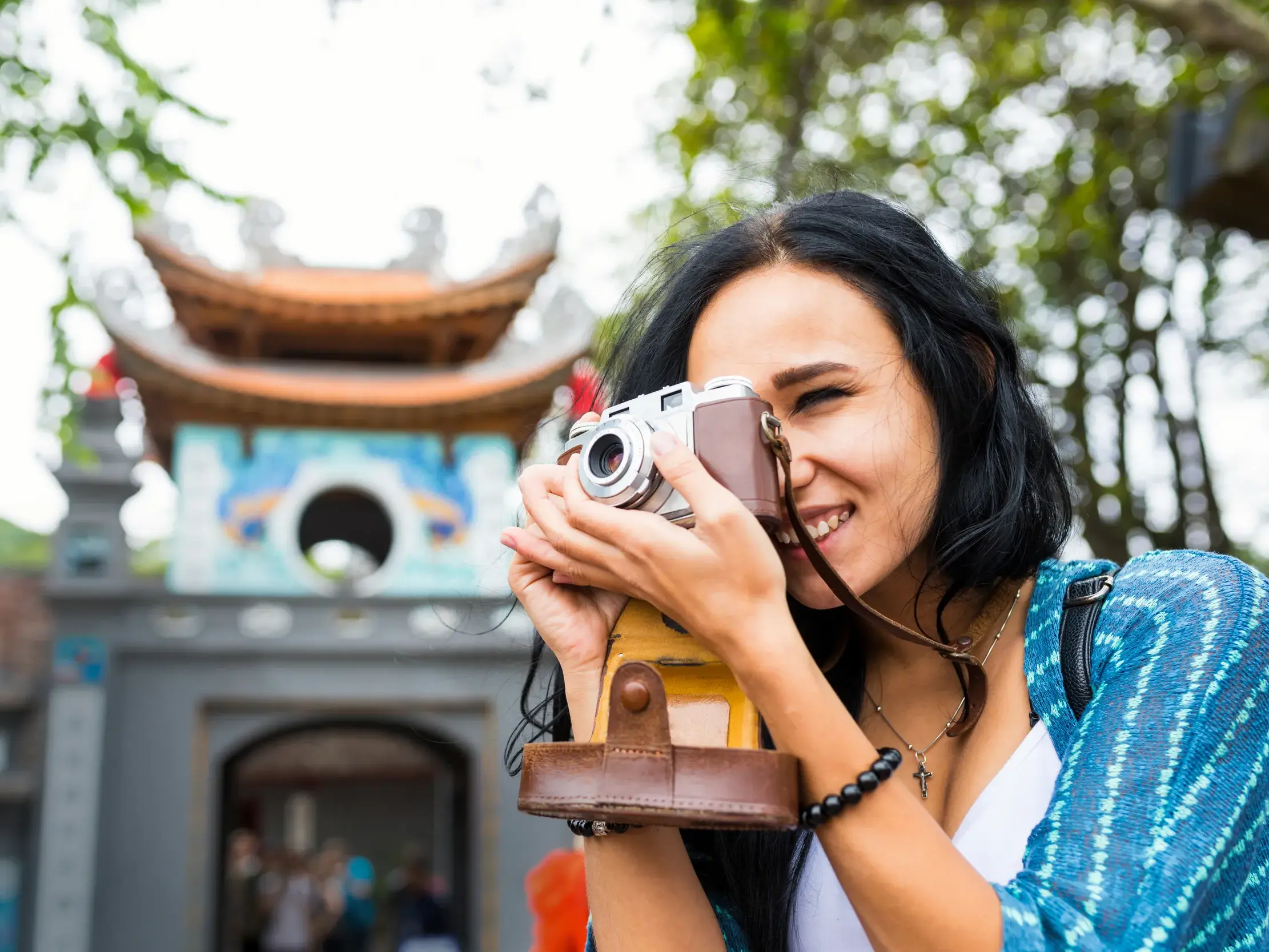 Eine junge Frau fotografiert lächelnd mit einer Retro-Kamera den Eingang zum Literaturtempel in Hanoi, Vietnam.