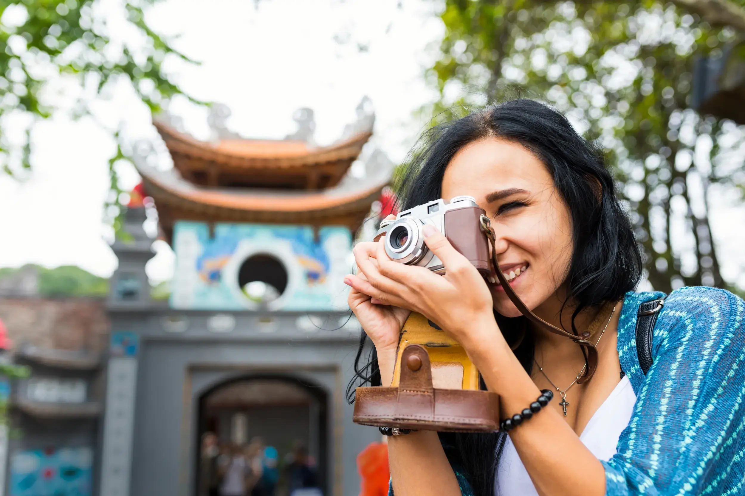Eine junge Frau fotografiert lächelnd mit einer Retro-Kamera den Eingang zum Literaturtempel in Hanoi, Vietnam.