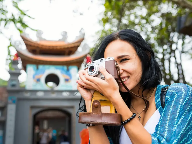 Eine junge Frau fotografiert lächelnd mit einer Retro-Kamera den Eingang zum Literaturtempel in Hanoi, Vietnam.