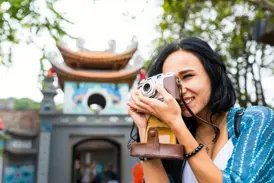 Eine junge Frau fotografiert lächelnd mit einer Retro-Kamera den Eingang zum Literaturtempel in Hanoi, Vietnam.