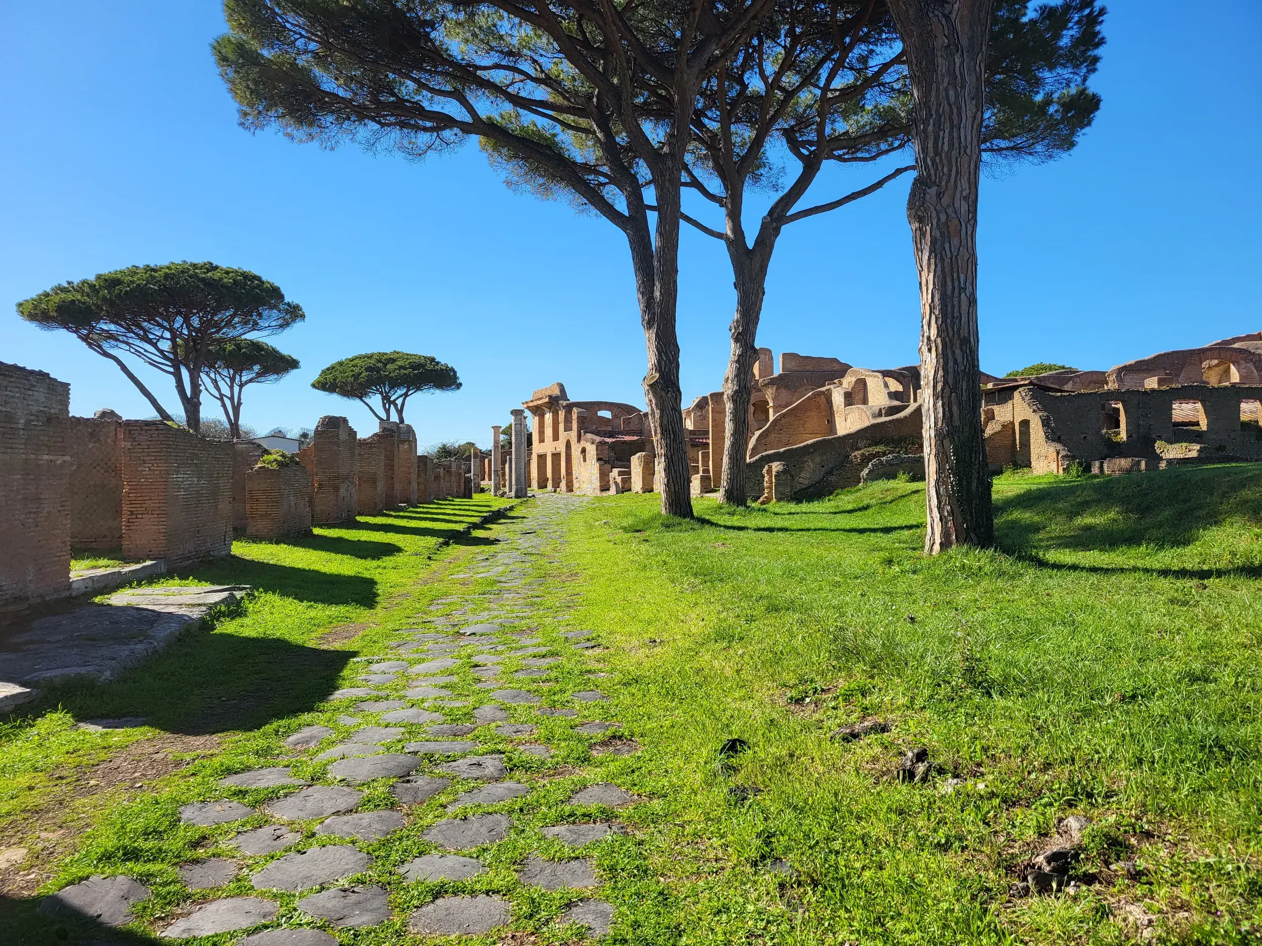 Ein alter Römerweg führt durch die sonnigen, grasbewachsenen Ruinen der antiken Hafenstadt Ostia Antica bei Rom.