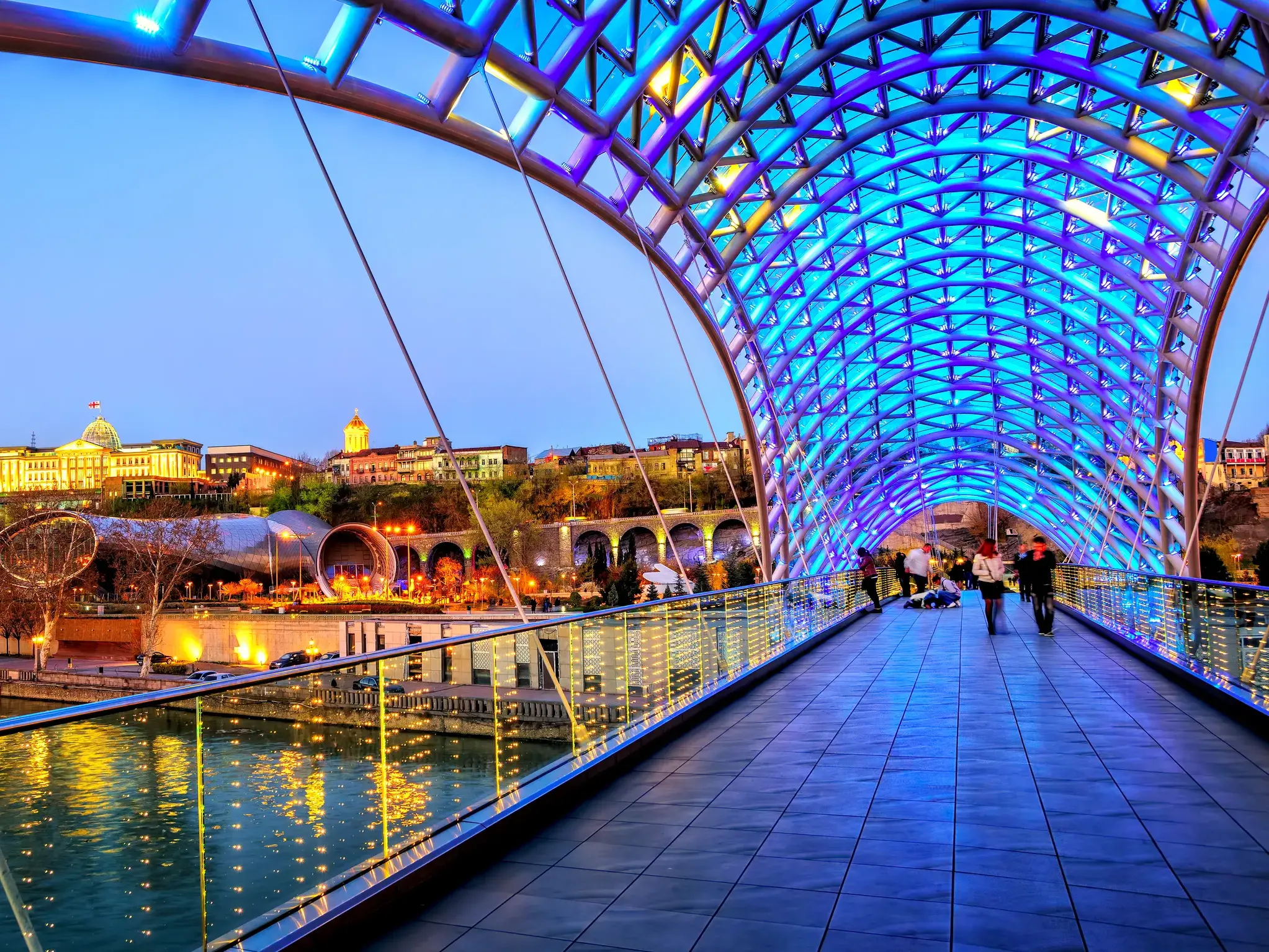 Die moderne Friedensbrücke in Tiflis leuchtet blau über dem Fluss Kura, im Hintergrund die historische Altstadt Georgiens.
