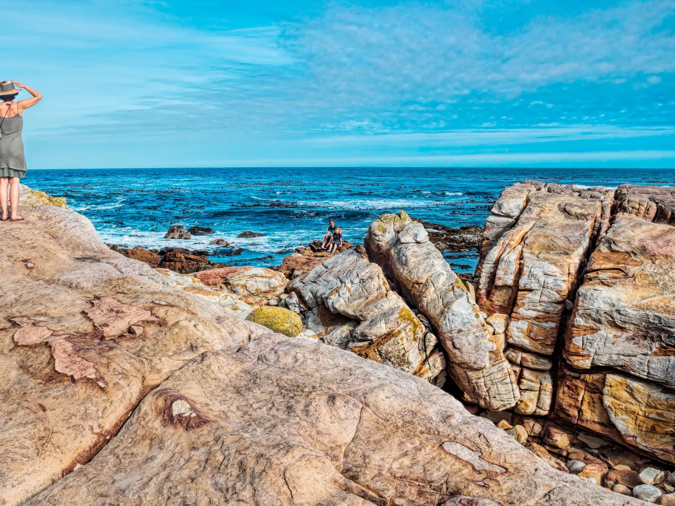 Eine Reisende mit Hut genießt den Ausblick von der Felsenküste bei Hermanus auf den weiten, blauen Ozean in Südafrika.