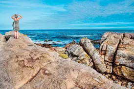 Eine Reisende mit Hut genießt den Ausblick von der Felsenküste bei Hermanus auf den weiten, blauen Ozean in Südafrika.
