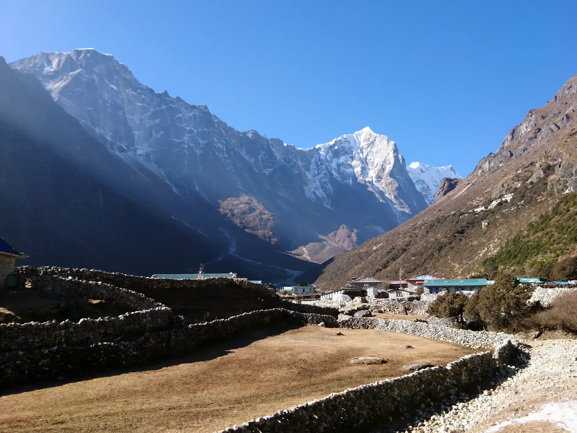 Blick über steingemauerte Weiden auf ein Dorf im Bhote Koshi-Tal mit schneebedeckten Gipfeln der Khumbu-Region in Nepal.