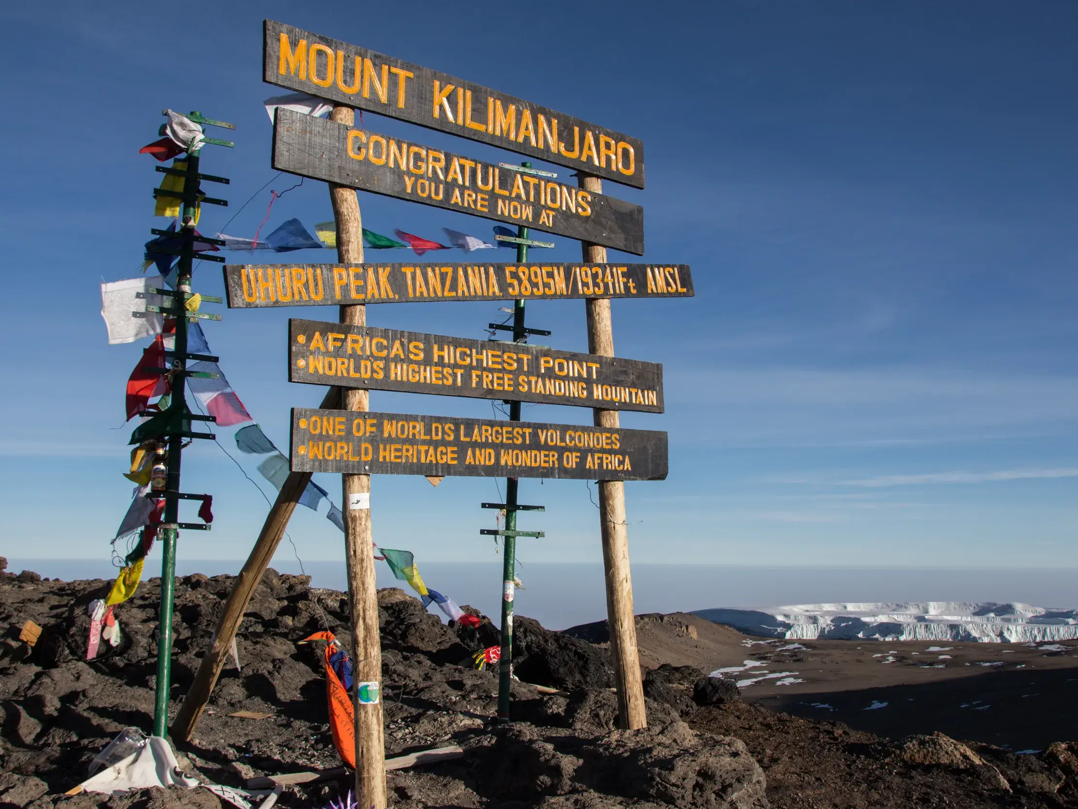 Das hölzerne Gipfelschild am Uhuru Peak auf dem Kilimanjaro, geschmückt mit Gebetsfahnen, vor dem schneebedeckten Kraterrand.