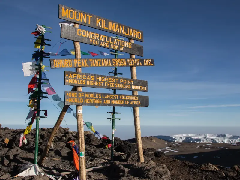 Das hölzerne Gipfelschild am Uhuru Peak auf dem Kilimanjaro, geschmückt mit Gebetsfahnen, vor dem schneebedeckten Kraterrand.