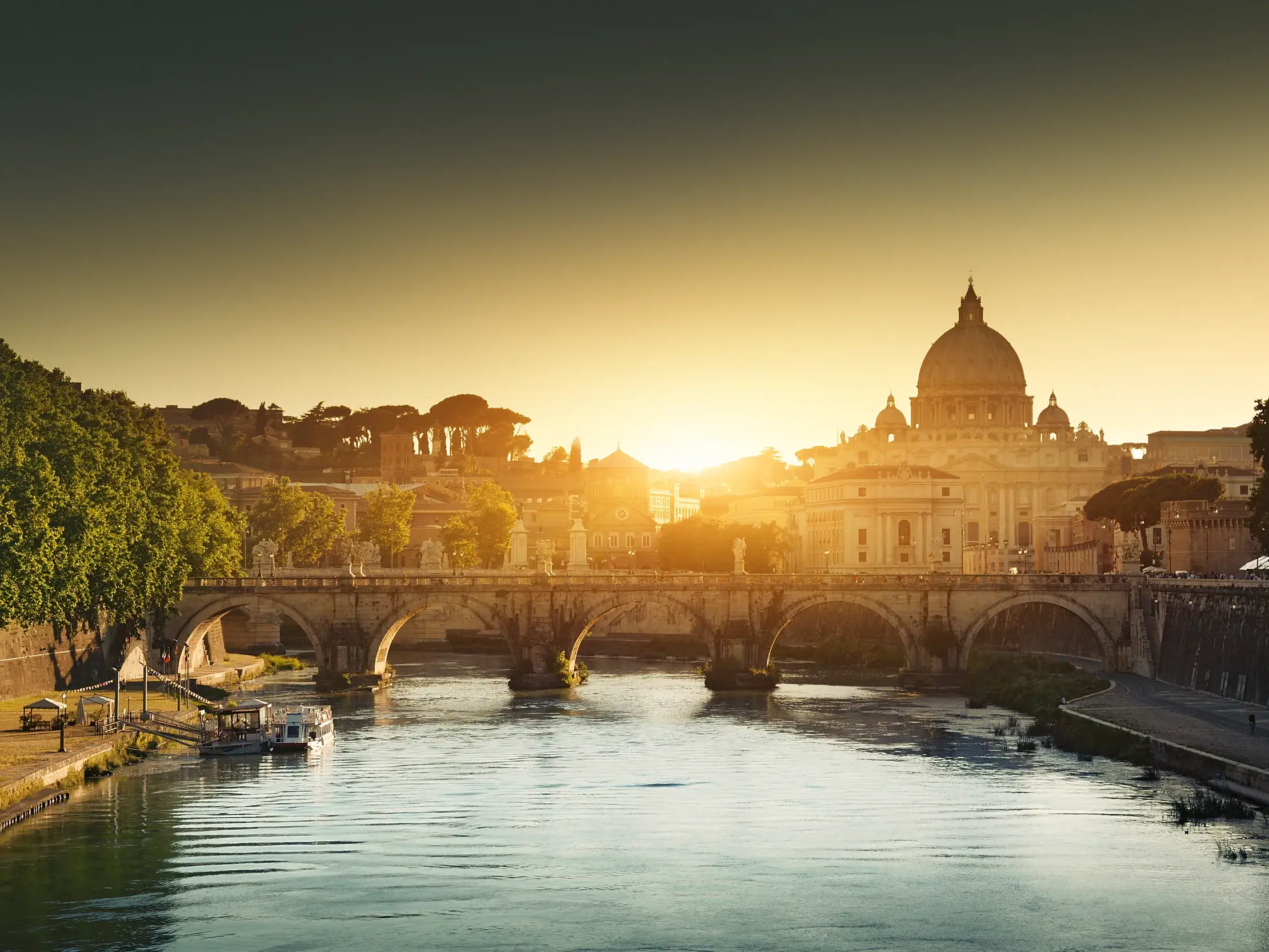 Blick über den Tiber auf eine historische Brücke und die Kuppel von St. Peter in Rom im warmen Sonnenlicht.
